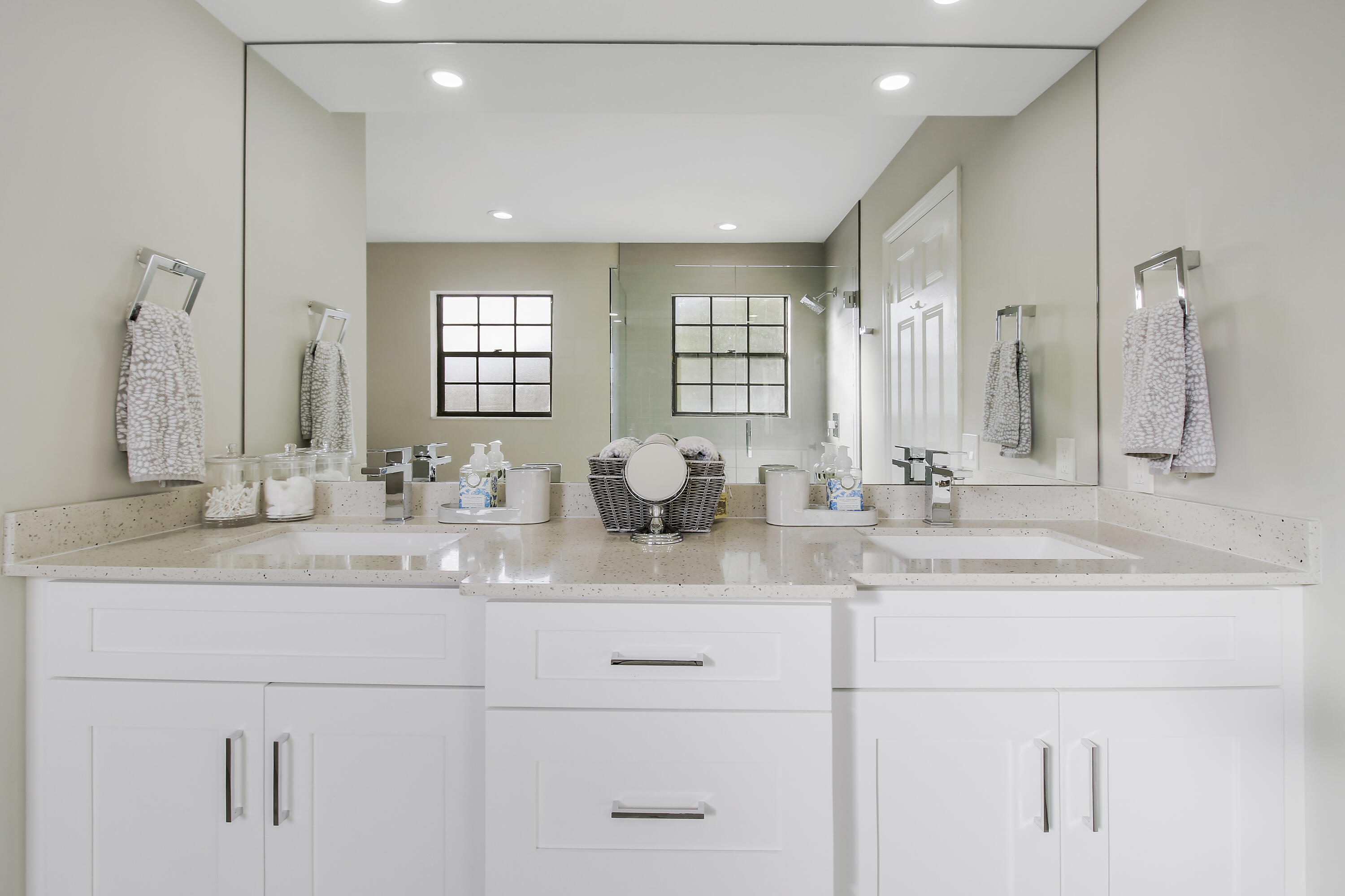 Bathroom vanity with two sinks, white cabinetry, and a mirrored wall reflecting the glass shower.