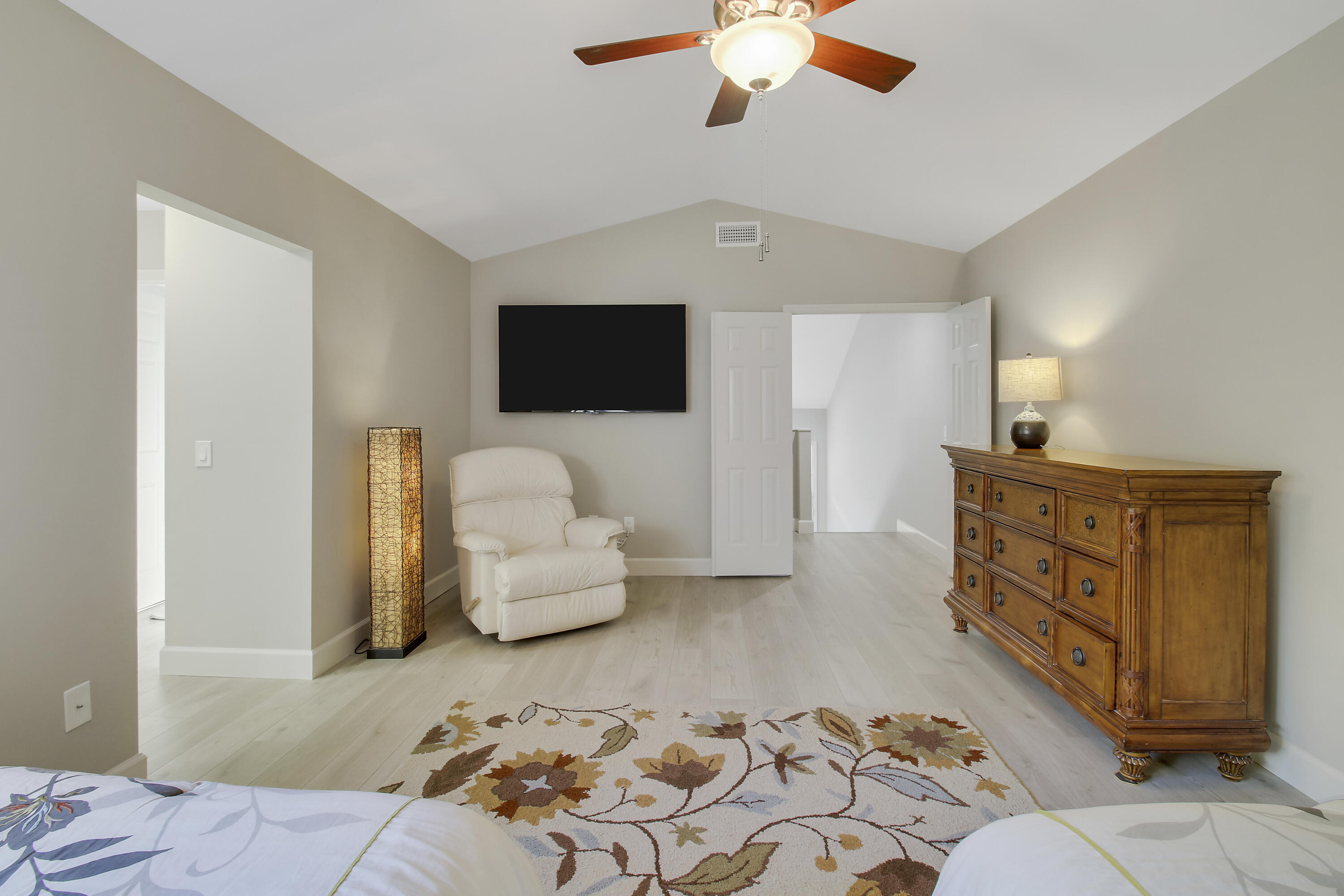 Furnished bedroom with a vaulted ceiling, wall-mounted TV, white recliner, and wooden dresser.