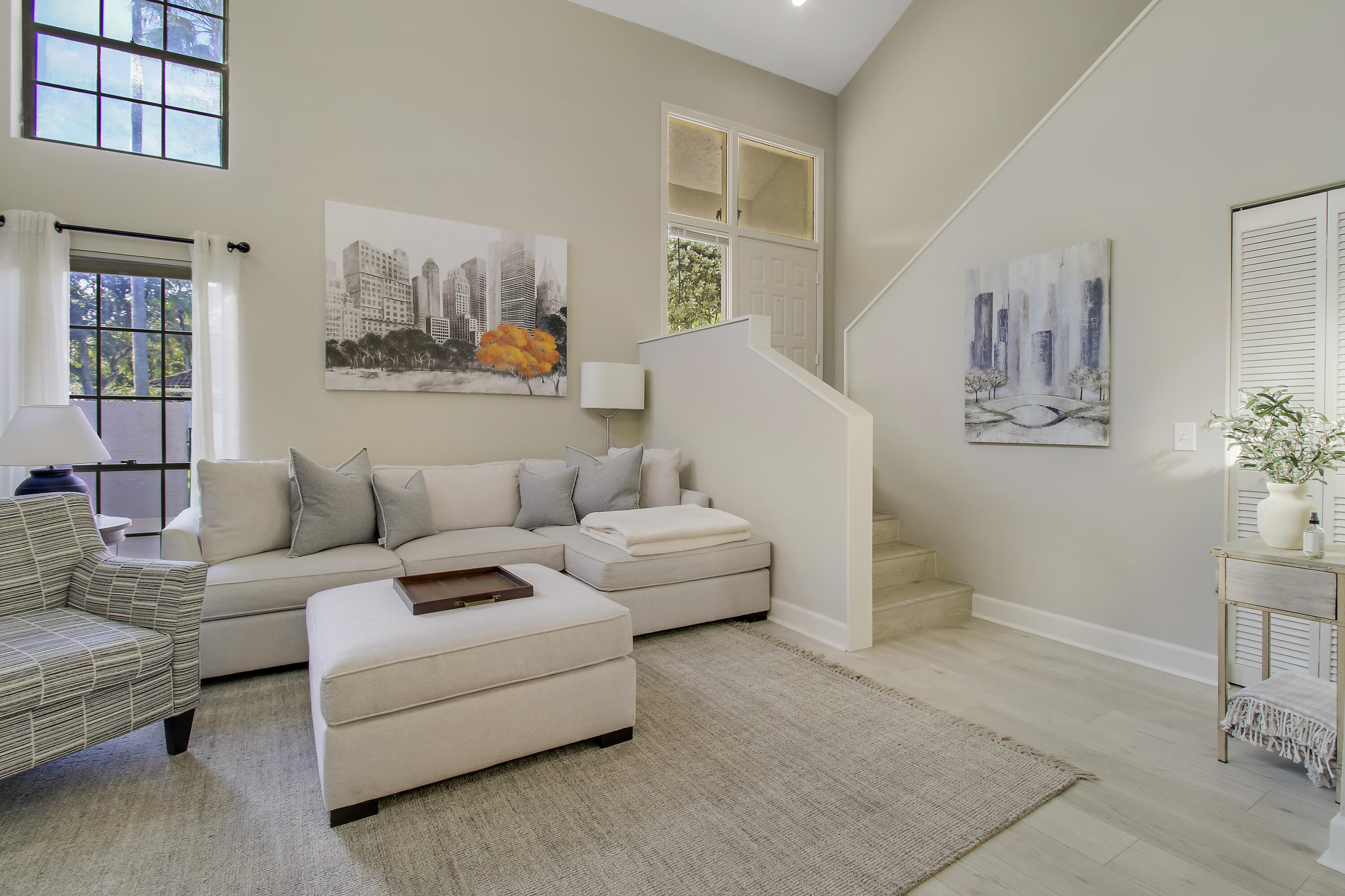 Living room with a light sectional sofa, woven armchair, tall windows, and staircase.