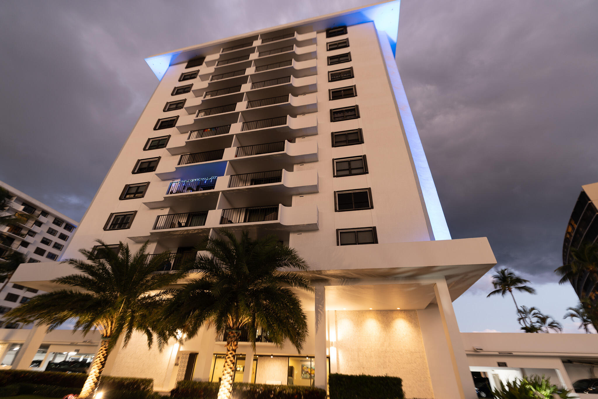 White condominium tower with stacked balconies and palm trees at dusk.