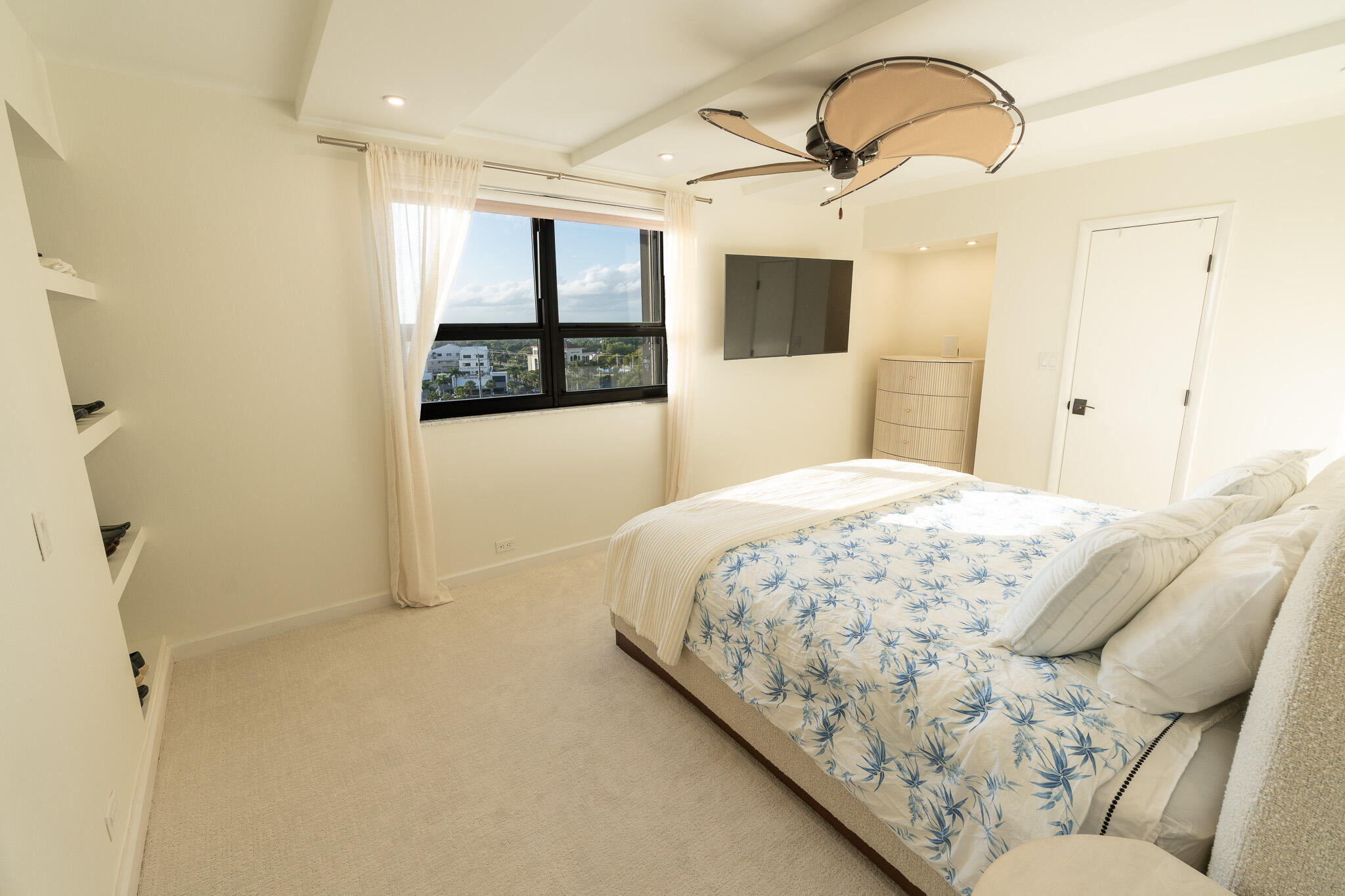 Bedroom with blue botanical bedding, a sculptural ceiling fan, and a large window overlooking nearby buildings.