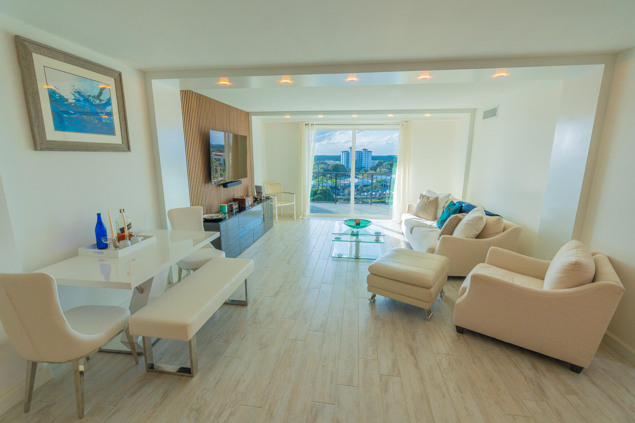 Furnished condo living room with cream seating and balcony doors overlooking buildings and greenery.