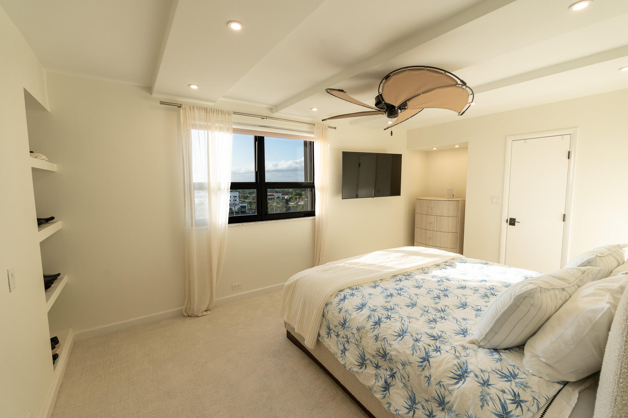 Bedroom with blue botanical bedding, a sculptural ceiling fan, and a window overlooking nearby buildings.