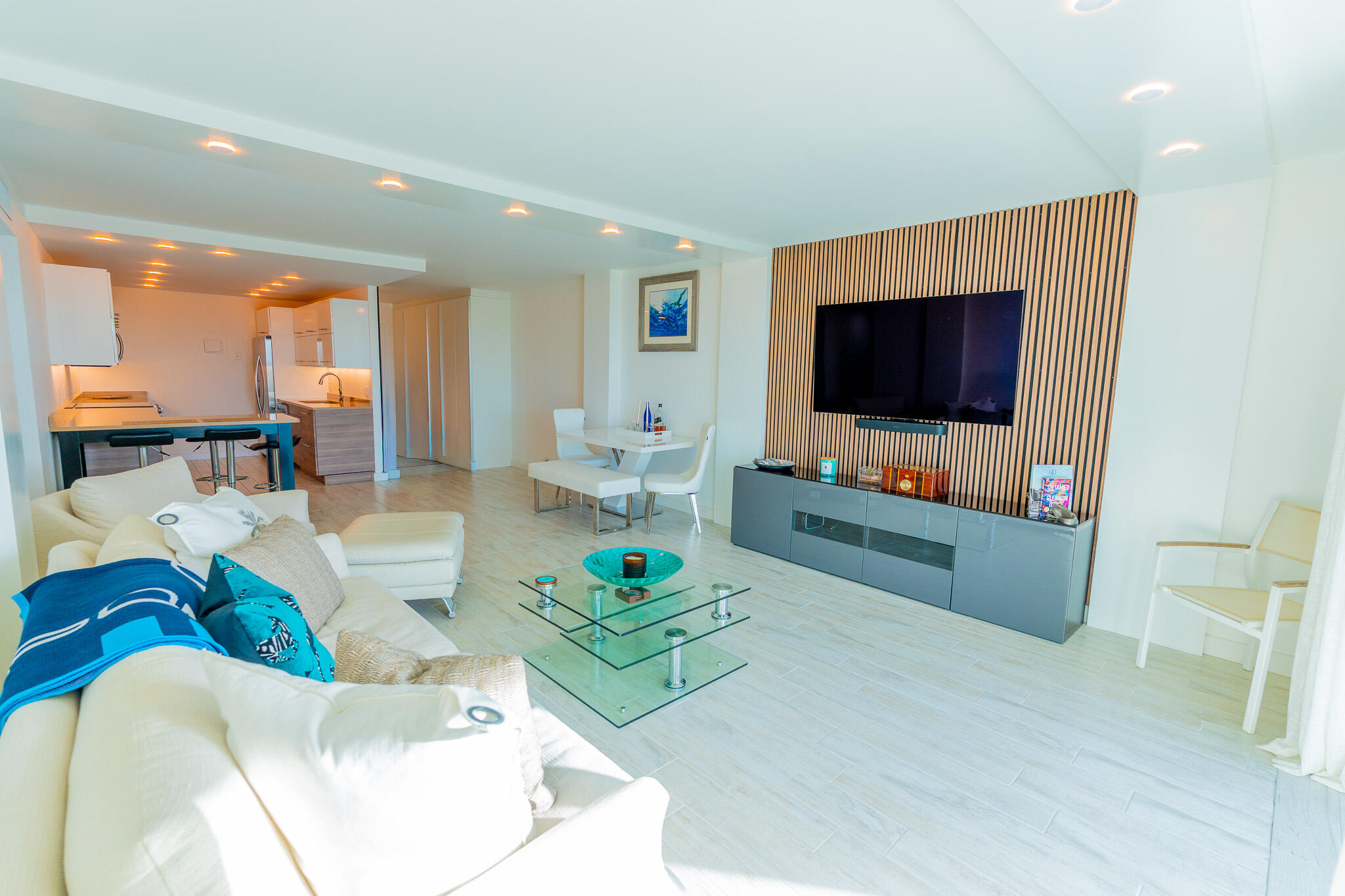 Open-plan living room with white sectional, glass coffee table, and television on a wood-slat wall.