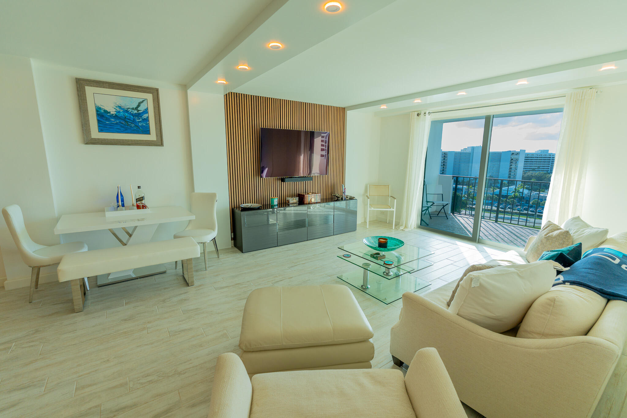 Condo living and dining area with white seating, a wall-mounted TV, and balcony views through sliding glass doors.
