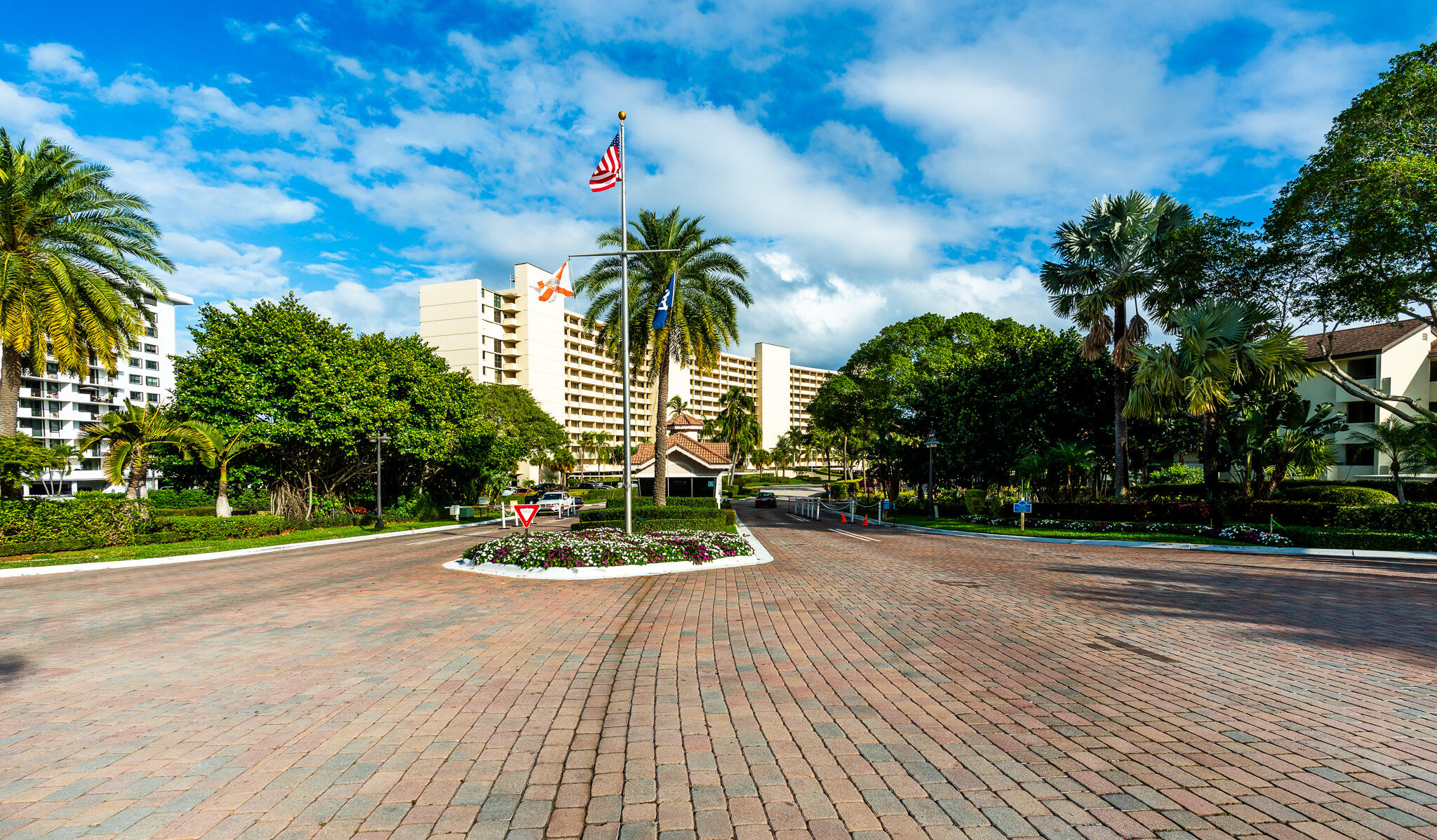 Condominium entrance with a brick-paved drive, tropical landscaping, and flagpoles.