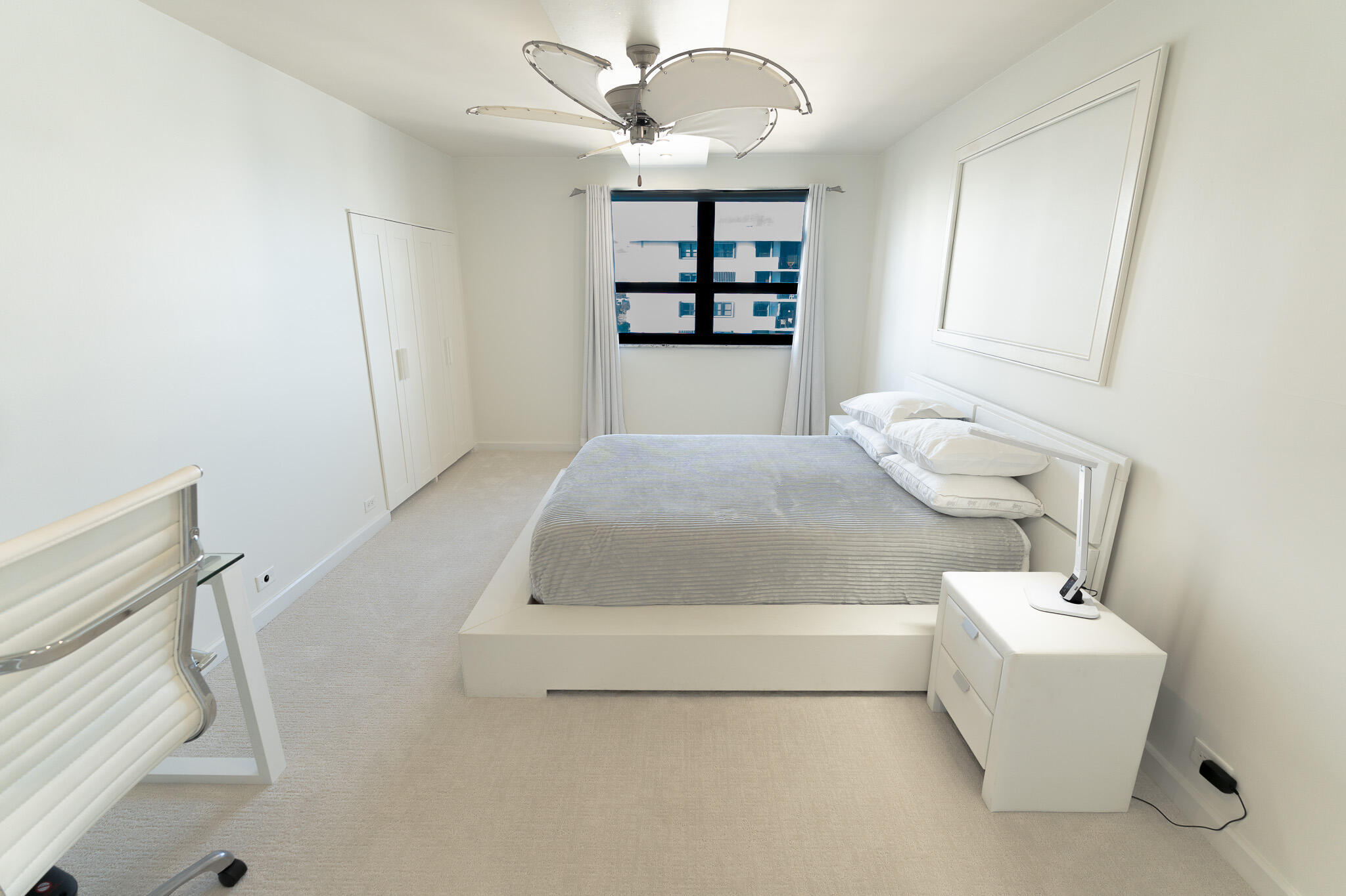 Bright bedroom with a low white bed, gray striped bedding, and a window overlooking a neighboring building.