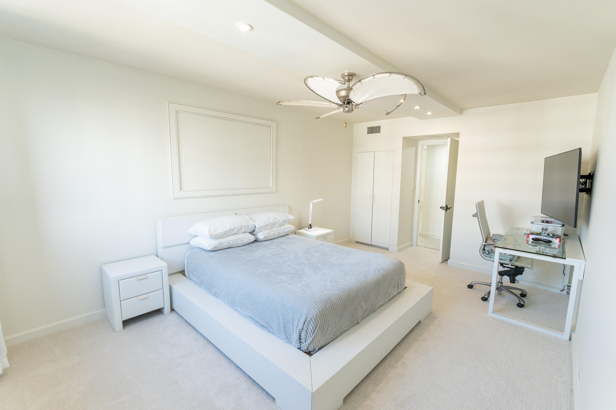 Bright bedroom with a pale blue platform bed, glass desk, wall-mounted television, and modern ceiling fan.