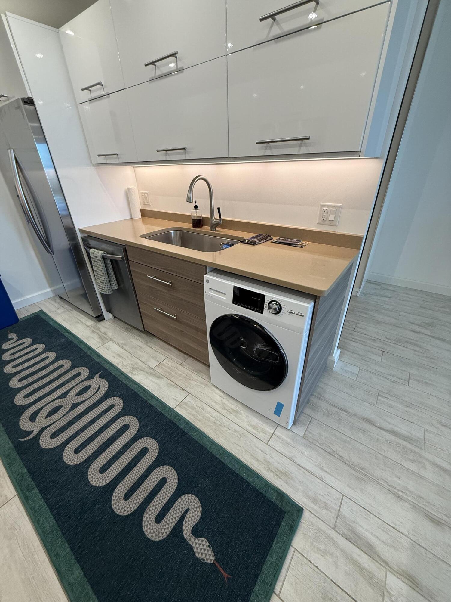 Laundry area with a front-loading washer, sink, refrigerator, and dark welcome mat.