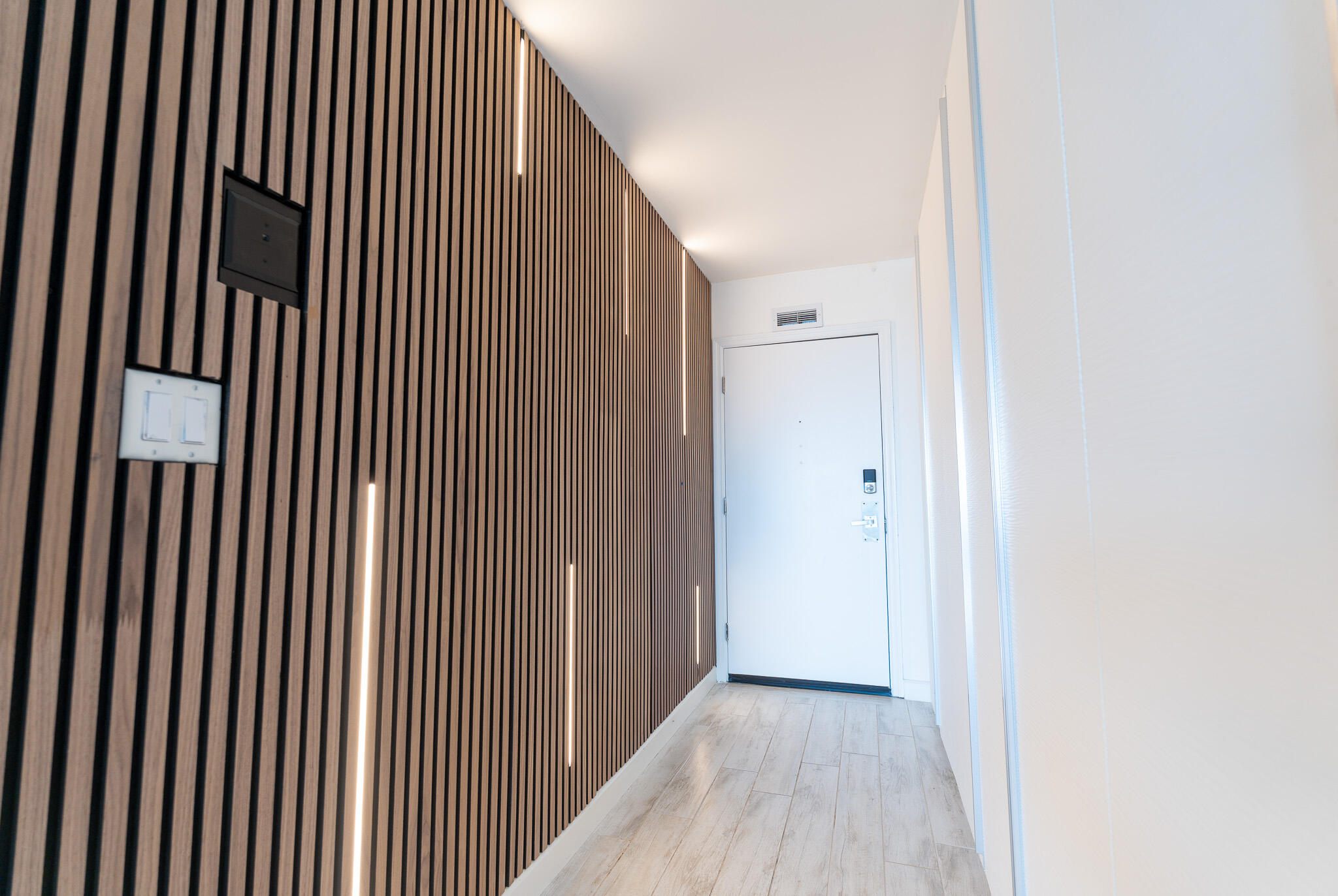 Apartment entry hallway with a lit wood-slat accent wall and pale tile flooring.