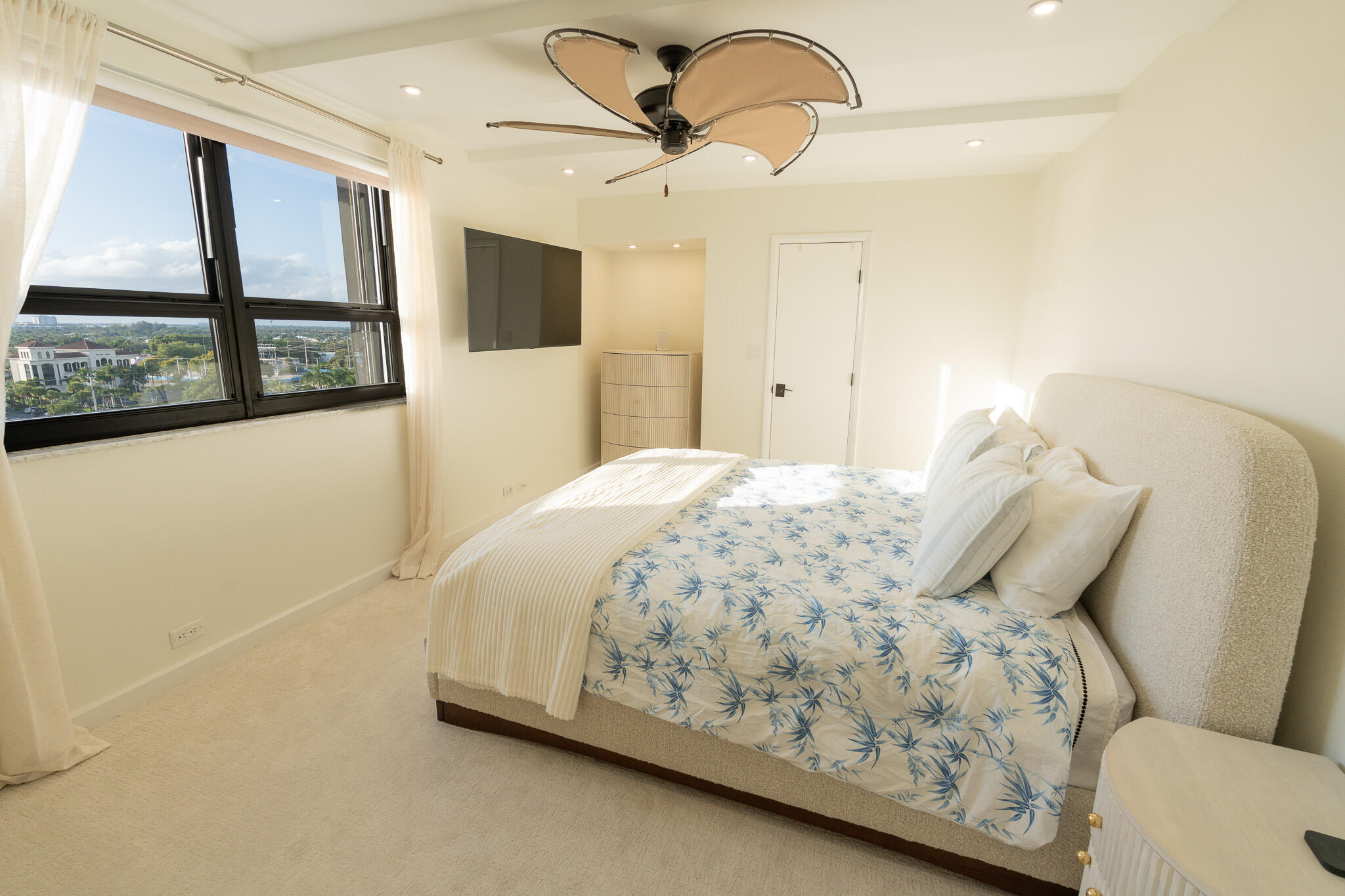 Bedroom with blue-patterned bedding, sculptural ceiling fan, and large windows overlooking the city.