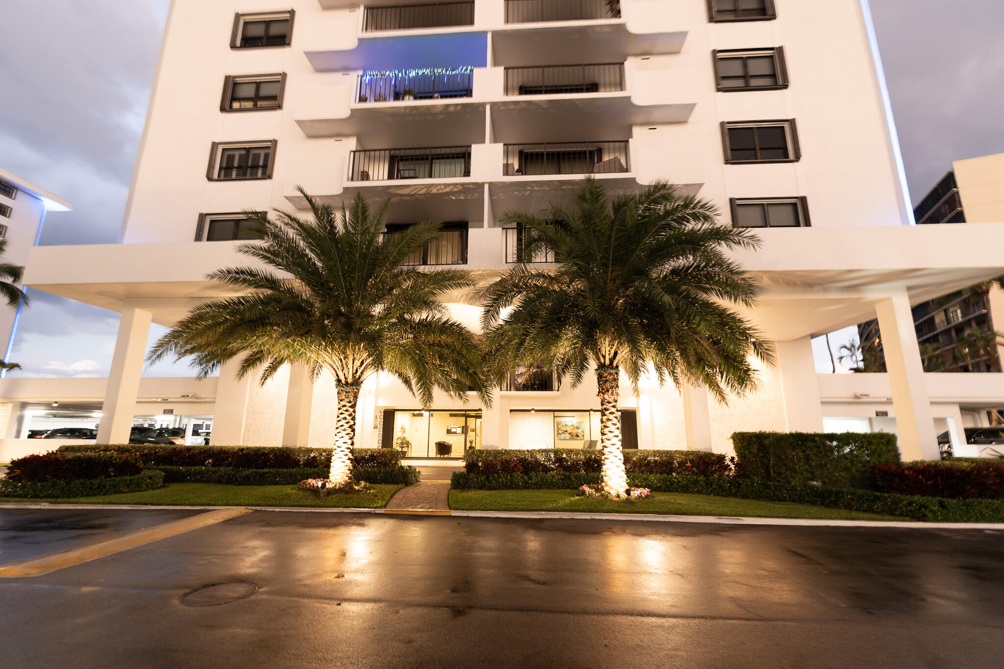 Condominium entrance at dusk, framed by two illuminated palm trees and a wet reflective driveway.