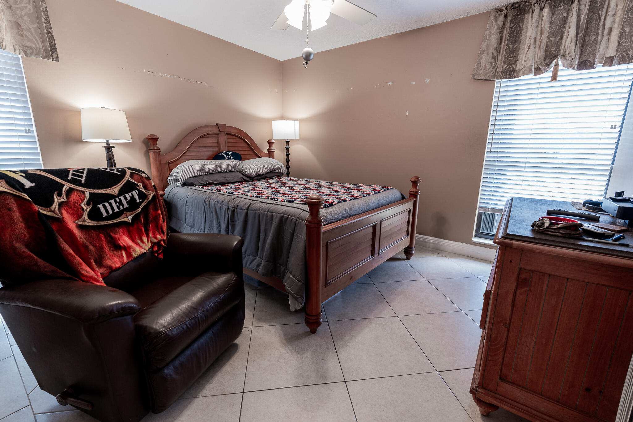 Bedroom with a wooden bed, leather recliner, tiled floor, and desk beside a window.