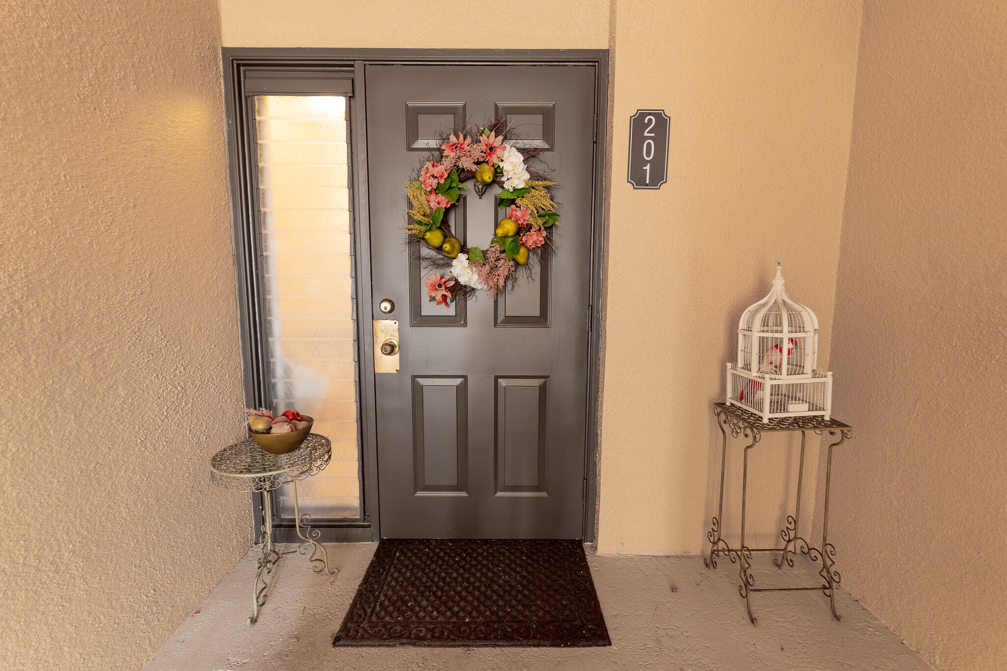 Unit 201’s dark front door with a colorful wreath, sidelight, and decorative birdcage.