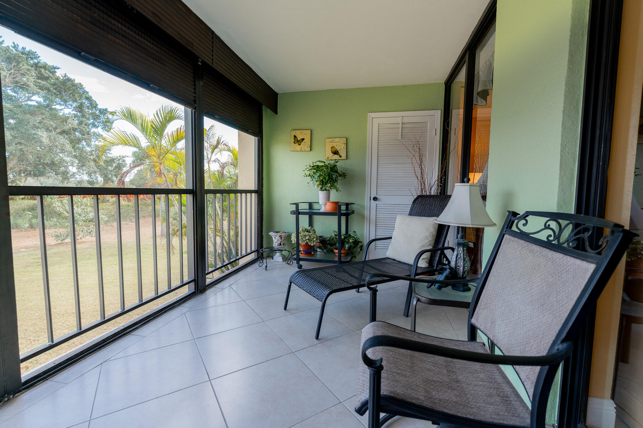 Screened balcony with black metal seating, potted plants, and a view of palms and lawn.