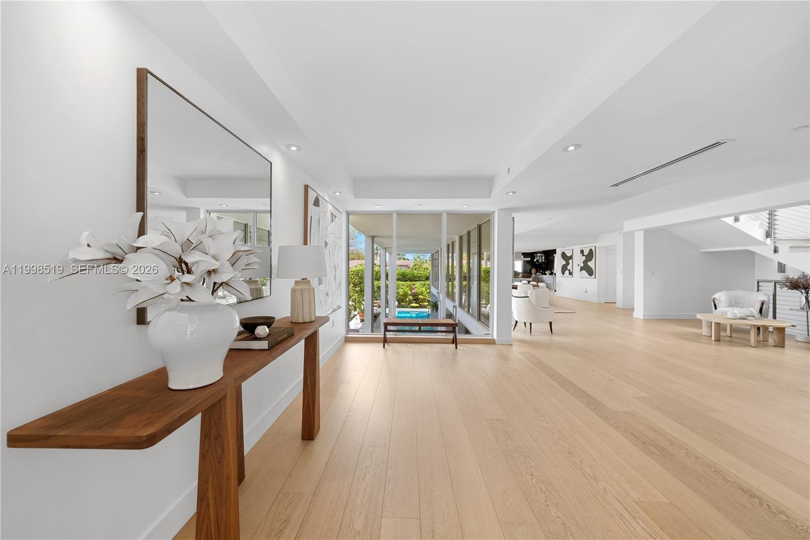 Modern open-plan interior with pale wood floors, a console table, and glass doors overlooking a pool.