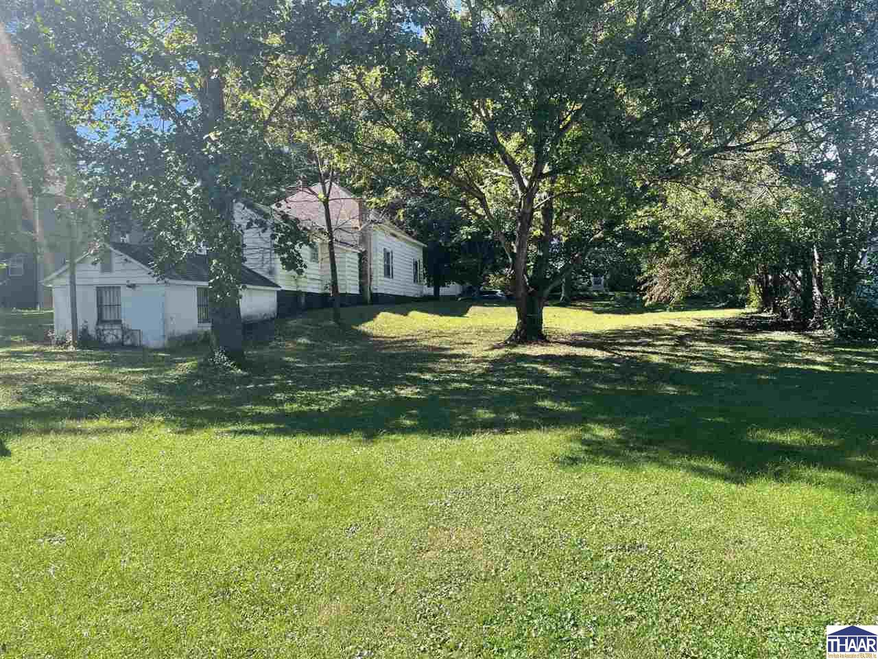 White-sided house beyond a broad lawn, shaded by mature trees, with a small outbuilding beside it.
