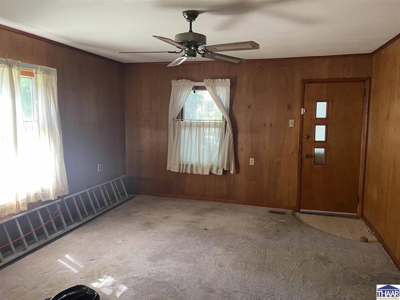 Empty carpeted room with wood-paneled walls, curtained windows, and a ceiling fan.