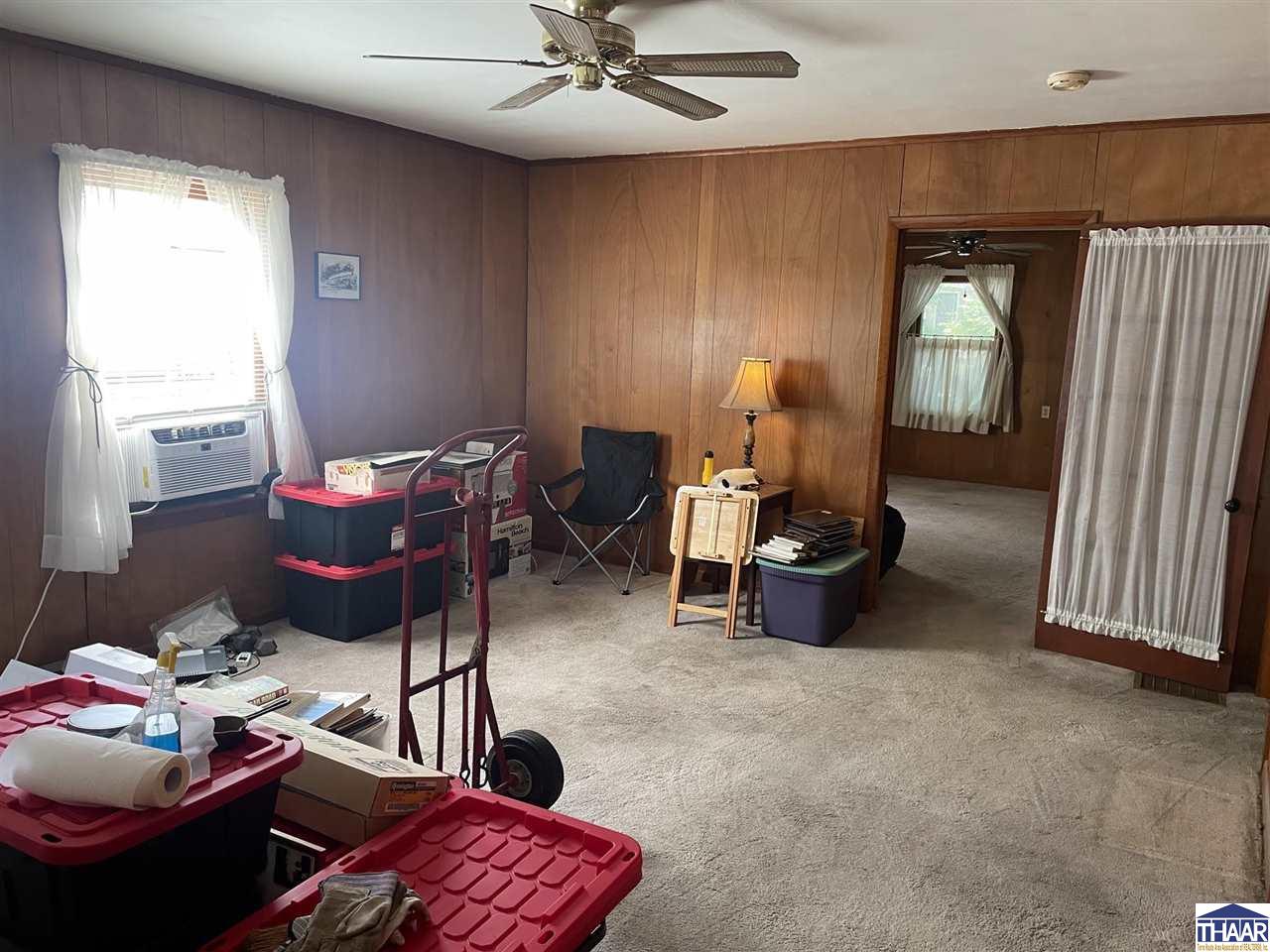 Carpeted room with wood-paneled walls, a ceiling fan, and a doorway to another room.