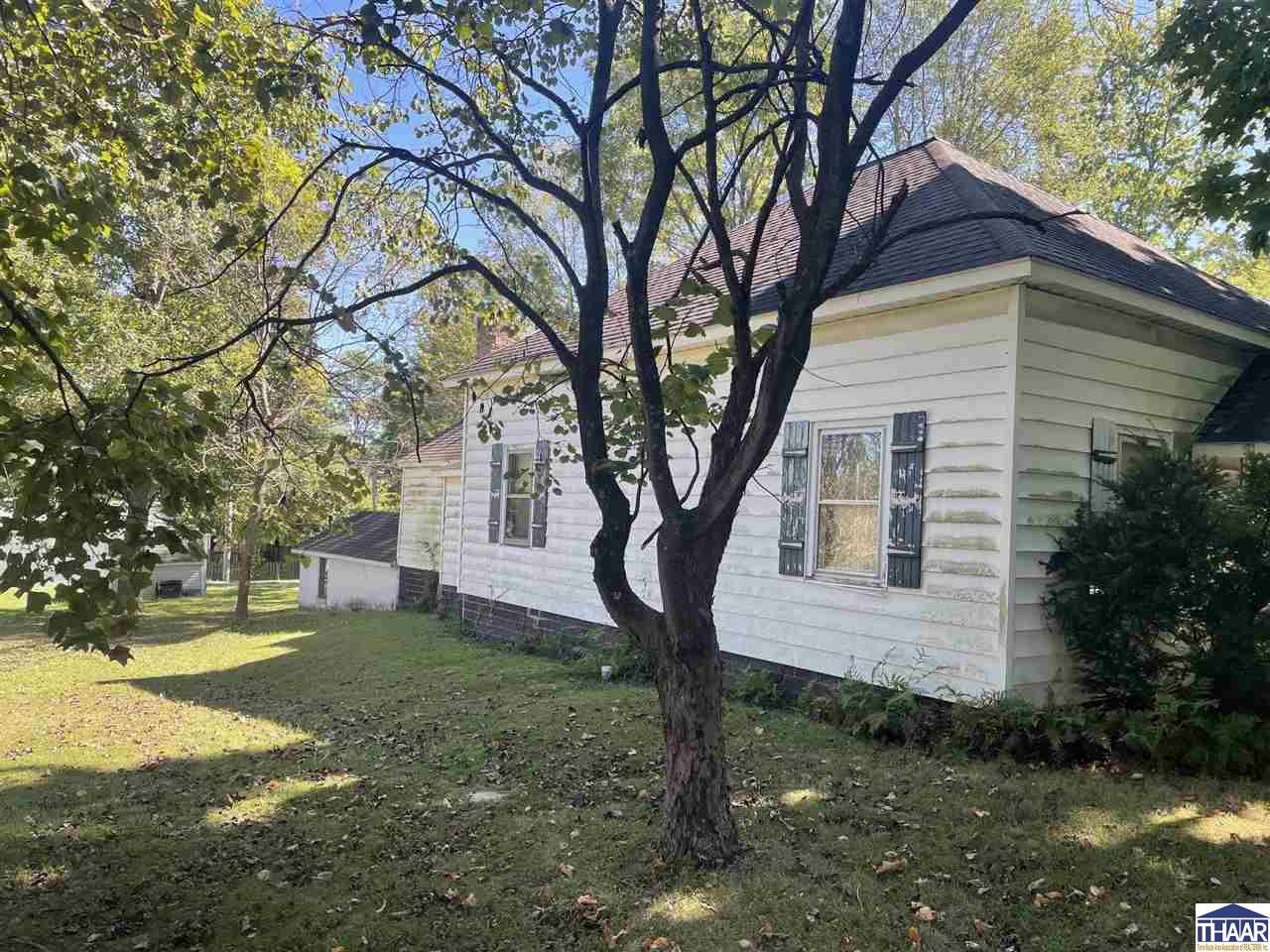 Light-colored two-story house partly screened by a large tree in a leafy yard.