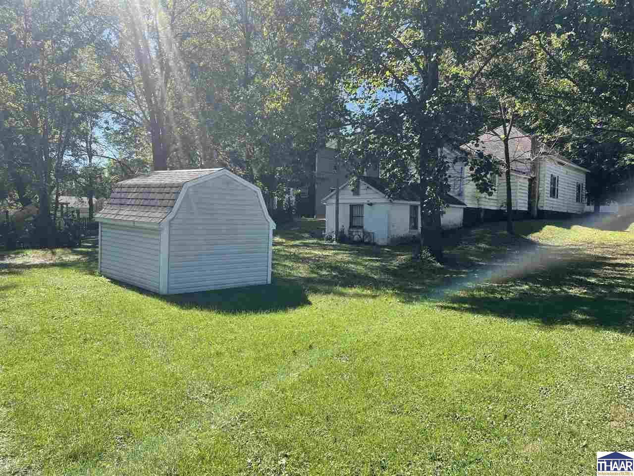 Grassy yard with a detached shed in front of a house beneath mature trees.