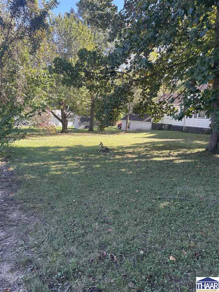 Residential yard with a broad lawn, mature shade trees, and part of a house in the background.
