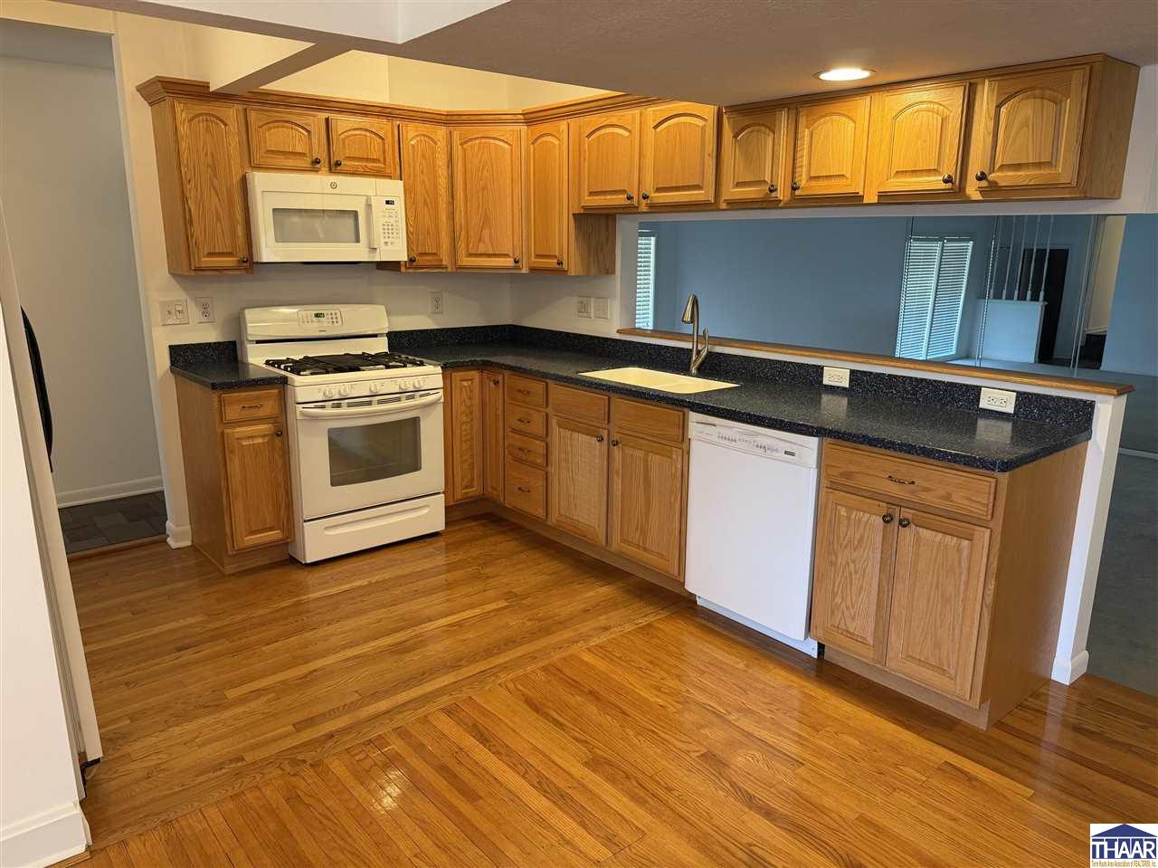 Kitchen with honey-colored wood cabinets, dark countertops, white appliances, and hardwood floors.