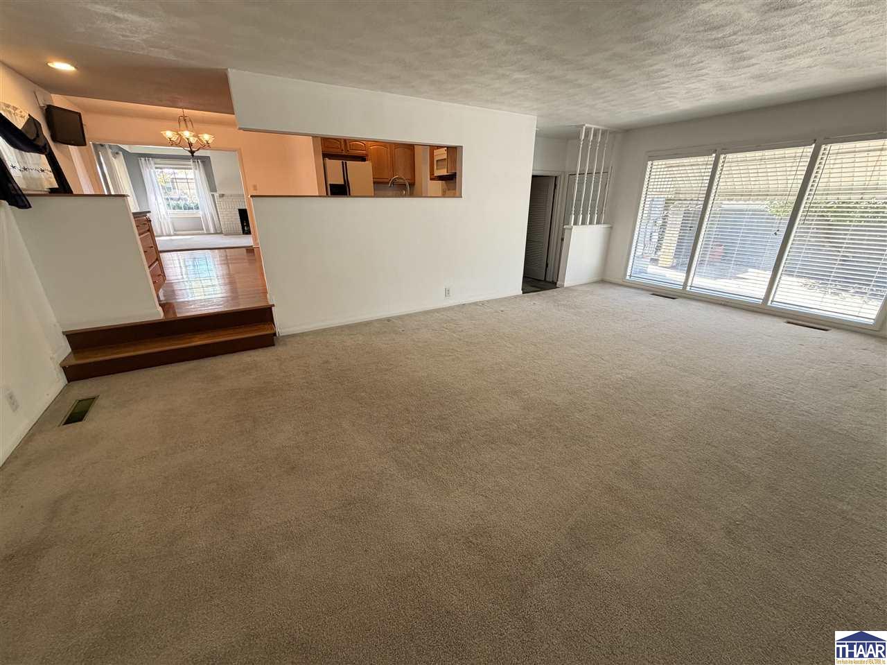 Empty carpeted living room with large windows and an open pass-through to the kitchen.