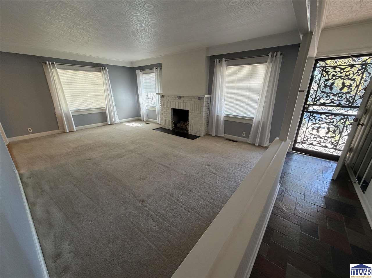 Empty living room with a white brick fireplace, patterned ceiling, and ornate glass entry door.