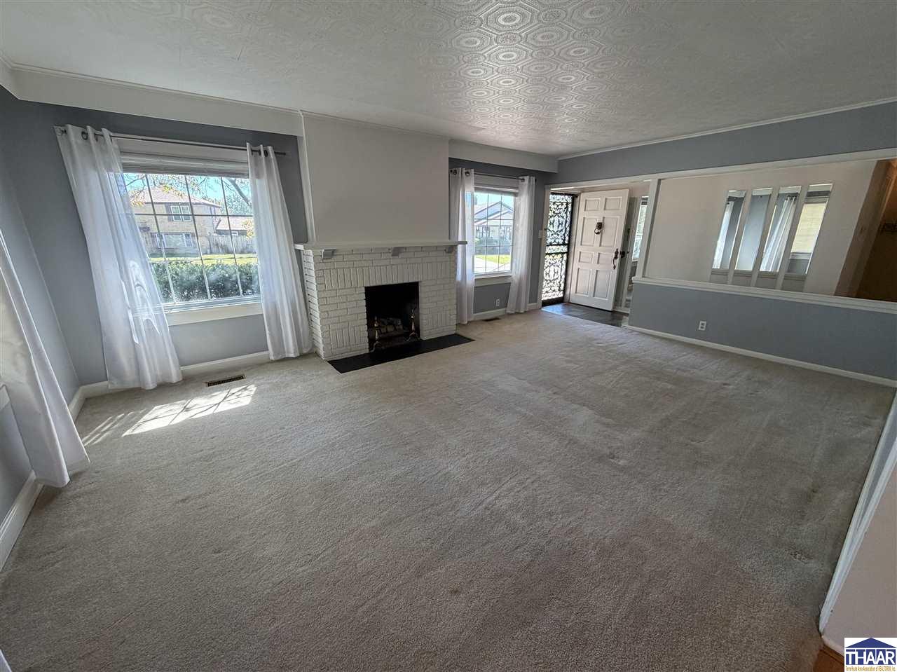 Empty living room with a white brick fireplace, large curtained windows, and a patterned ceiling.