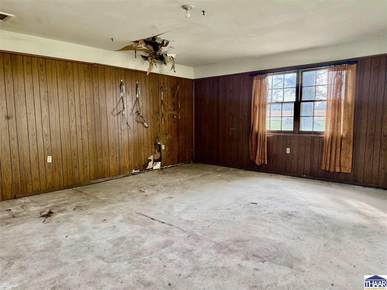 Vacant wood-paneled room with a damaged ceiling, damaged wall, concrete floor, and curtained window.