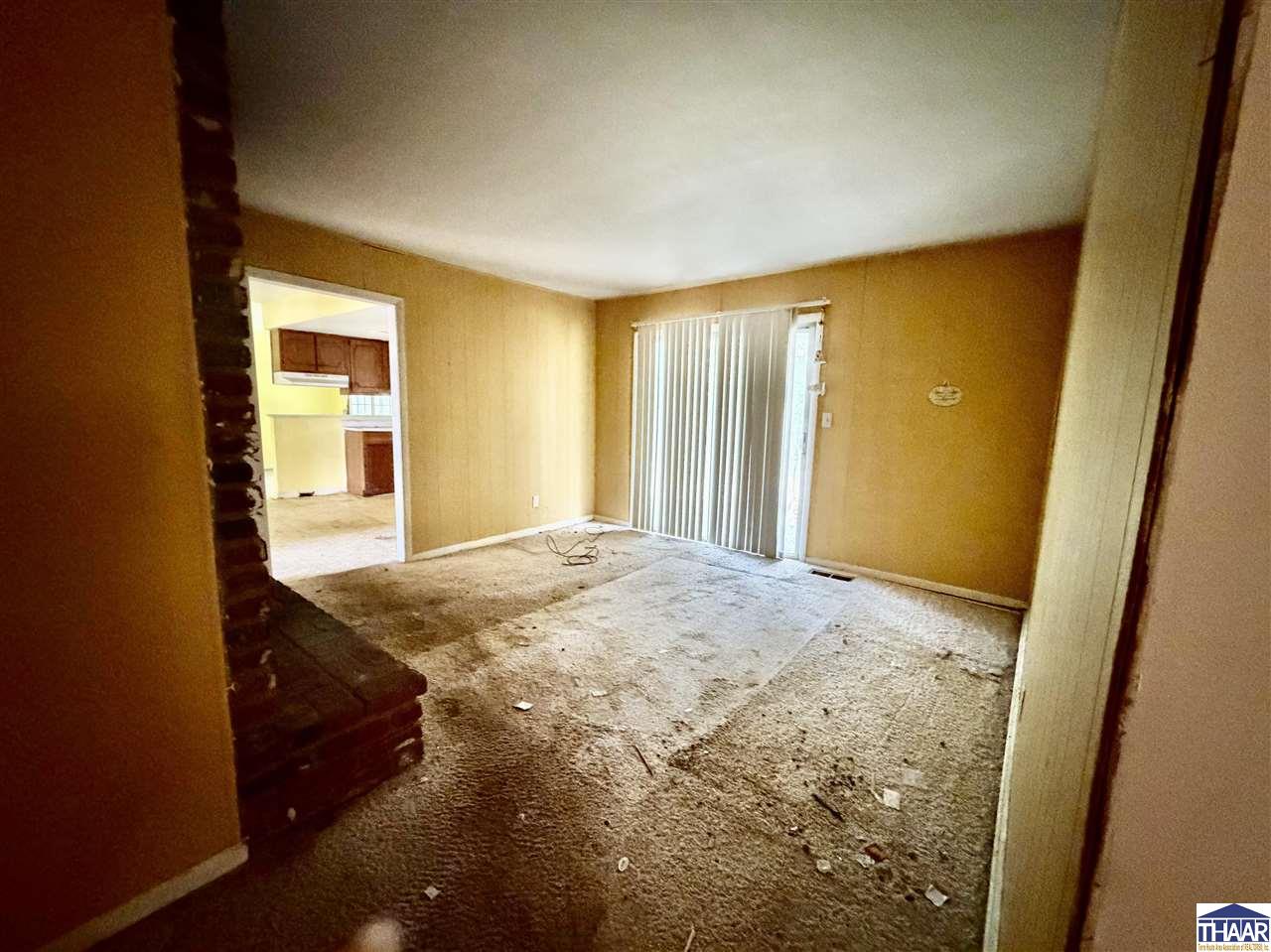 Vacant living room with stained carpet, a brick fireplace, and vertical blinds over a sliding glass door.