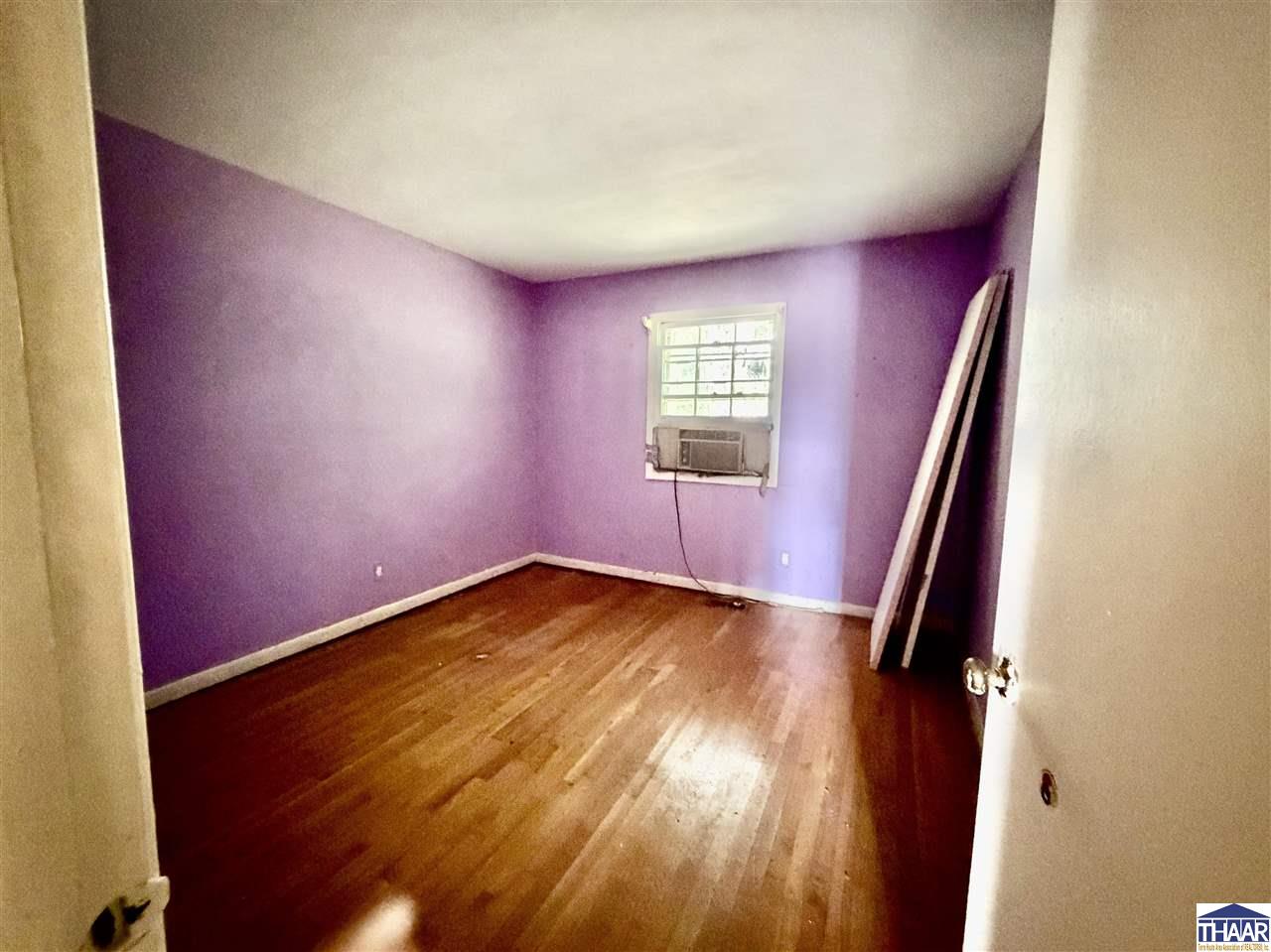 Empty purple-walled room with hardwood floors, a window air conditioner, and boards leaning by the window.