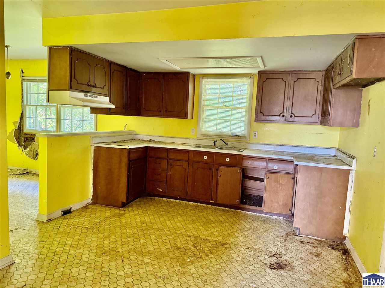 Vacant kitchen with yellow walls, dark wood cabinets, and damaged flooring and wall surfaces.
