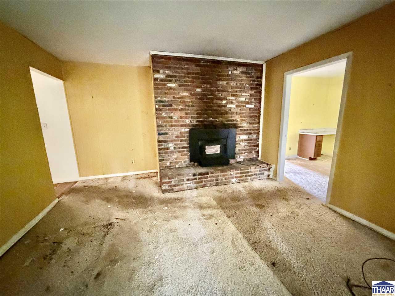 Empty living room with a brick fireplace and black stove insert, stained carpet, and an adjoining room visible through the doorway.
