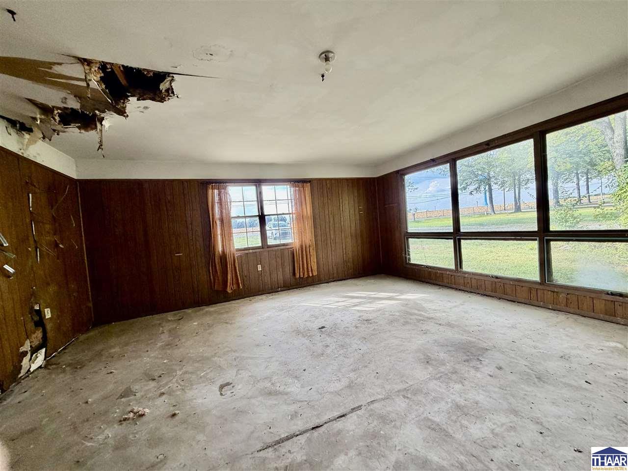 Empty wood-paneled living room with damaged ceiling and large windows overlooking water.
