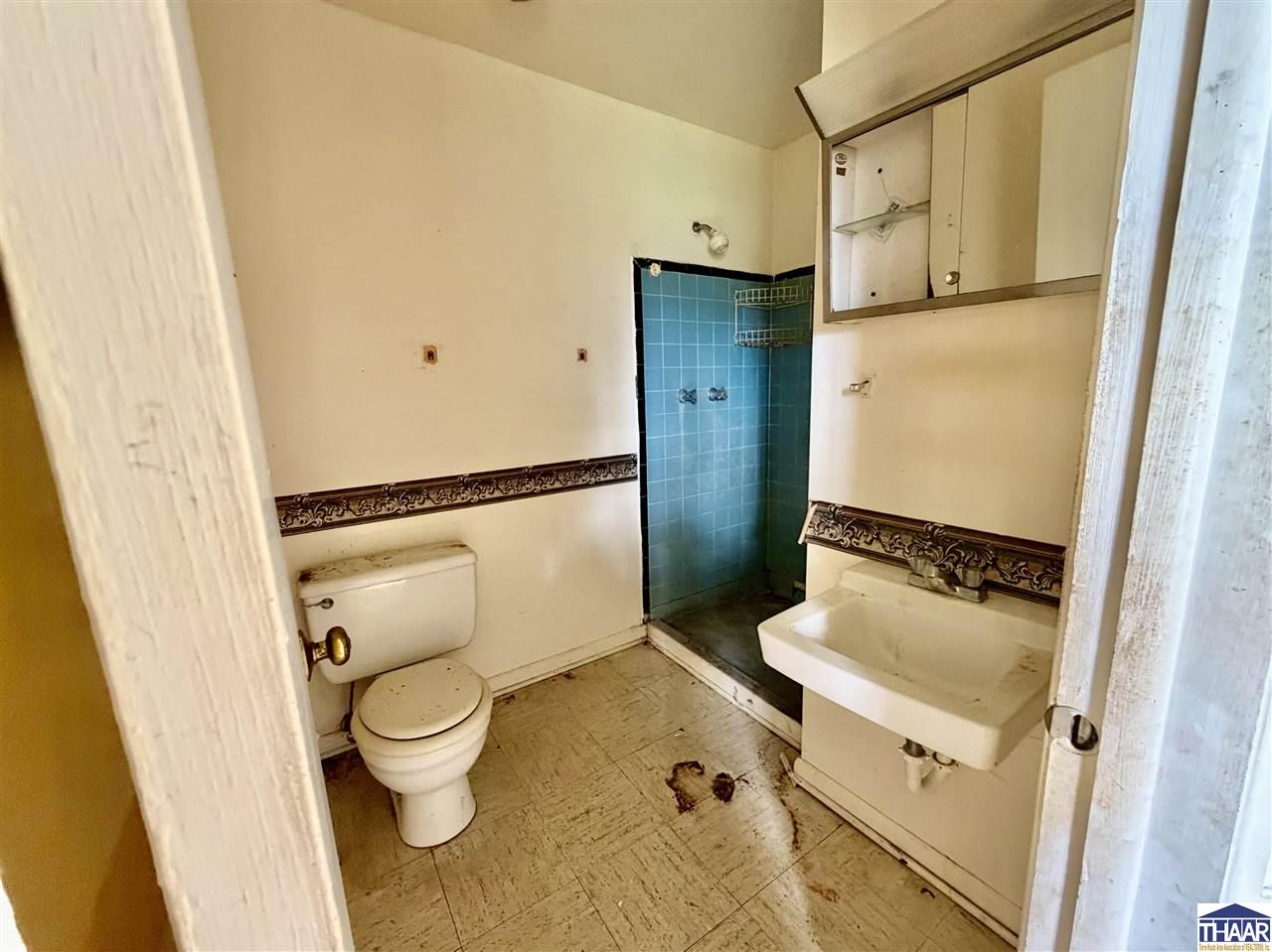 Older bathroom with a blue-tiled shower, toilet, wall-mounted sink, and patterned wall trim.
