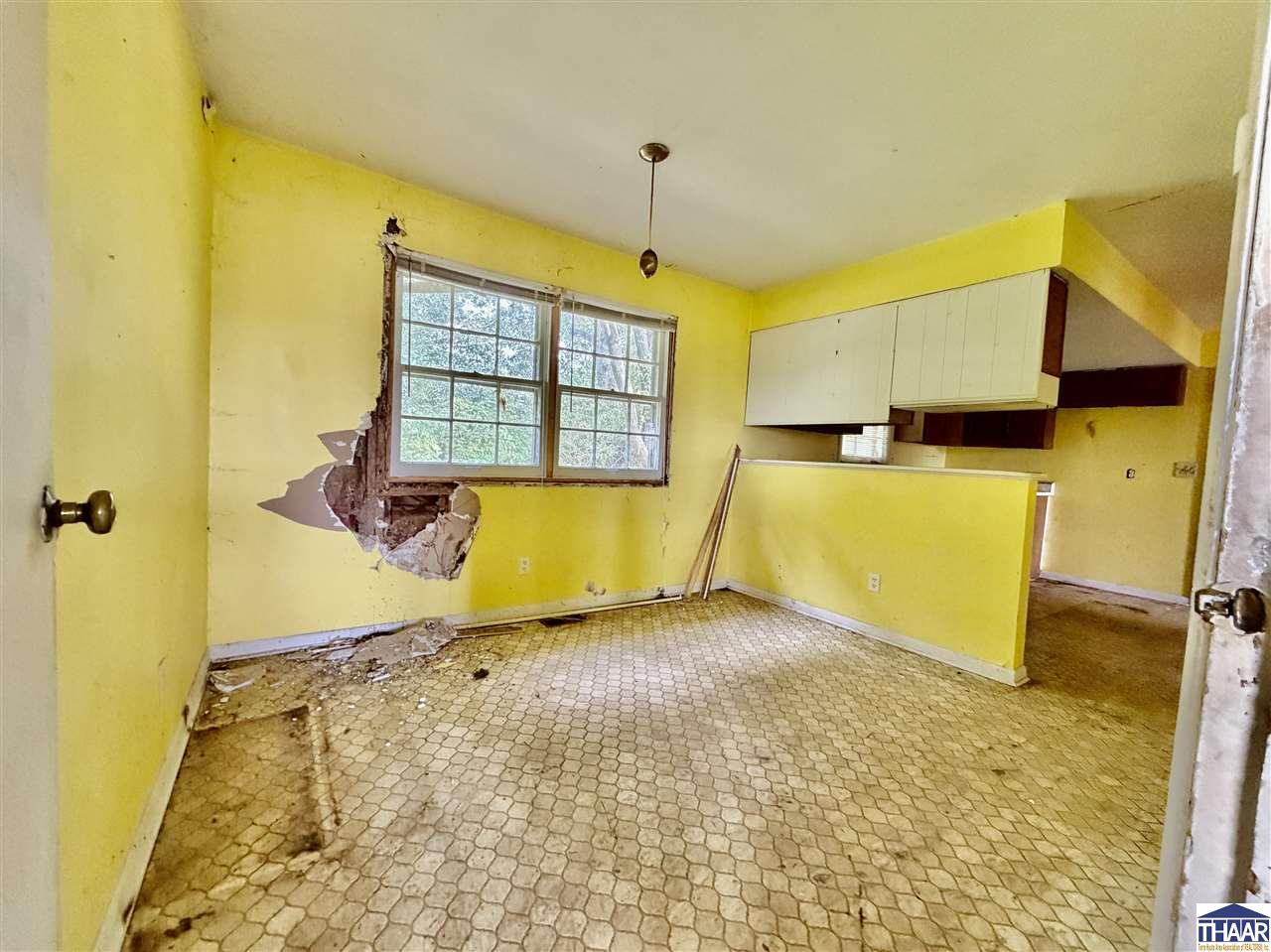 Vacant kitchen with yellow walls, damaged plaster below two windows, and worn patterned flooring.
