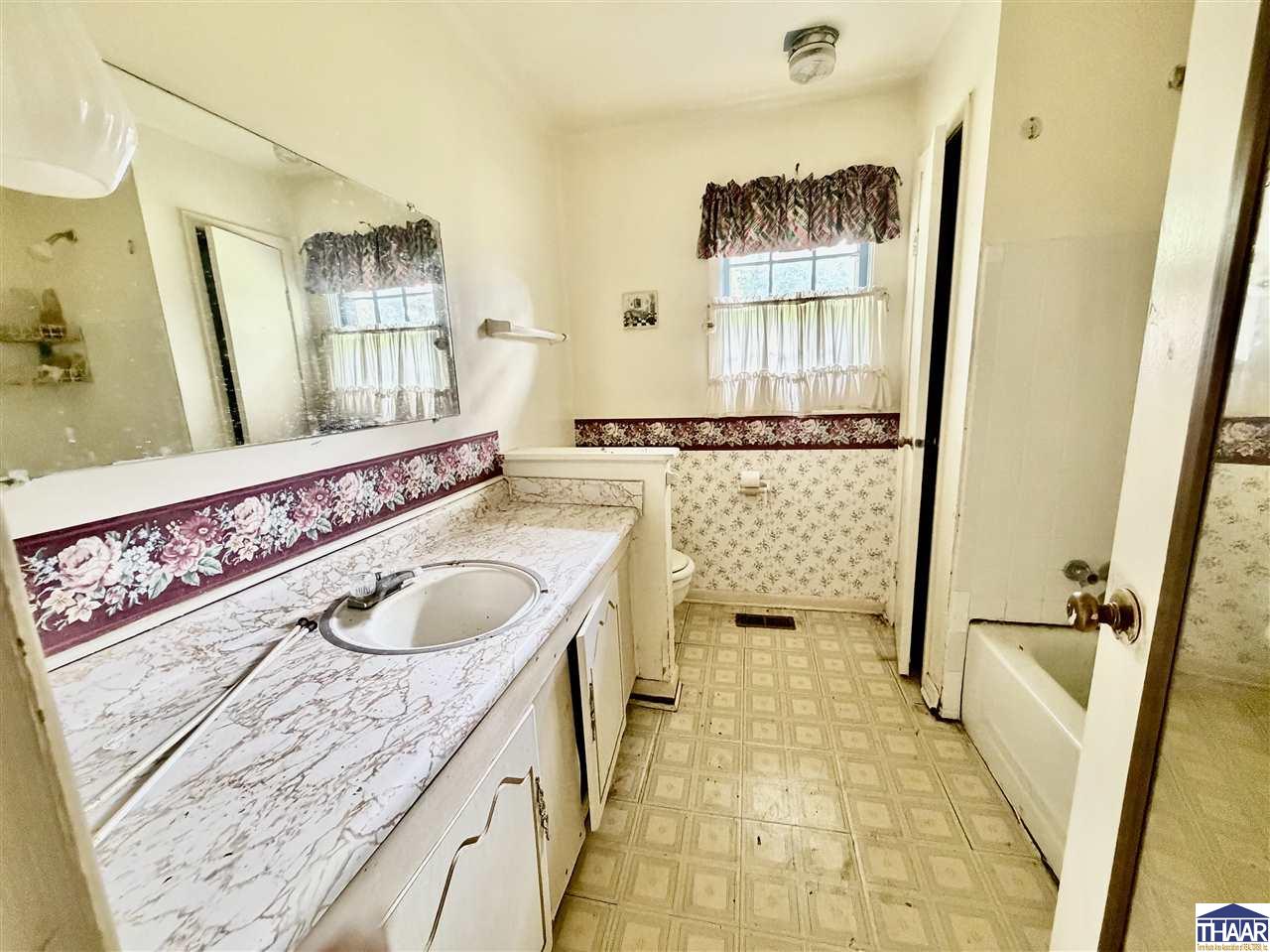 Bathroom with a long mirrored vanity, floral wallpaper, curtained window, and bathtub.