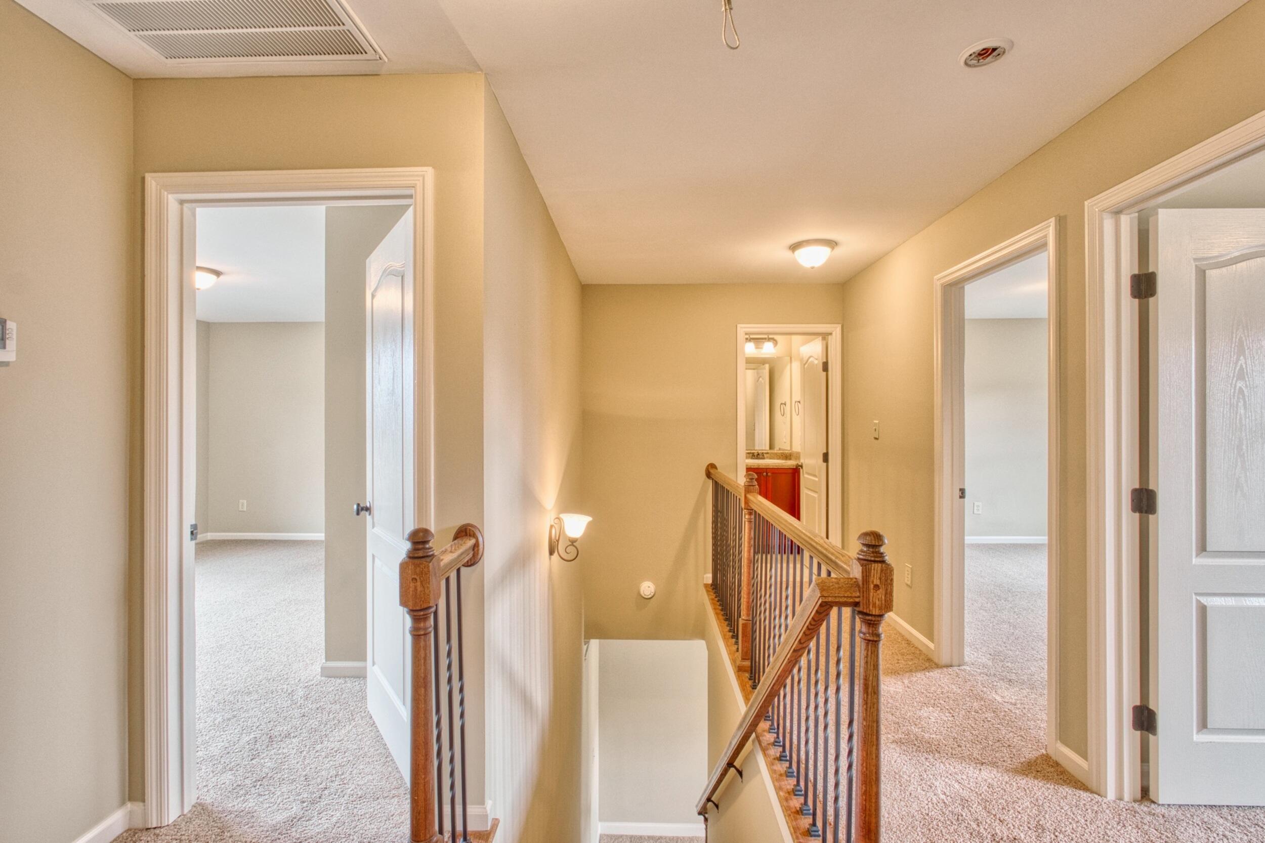 Upstairs hallway with carpeted floors and a wooden railing around the stairwell; open rooms and a bathroom are visible.