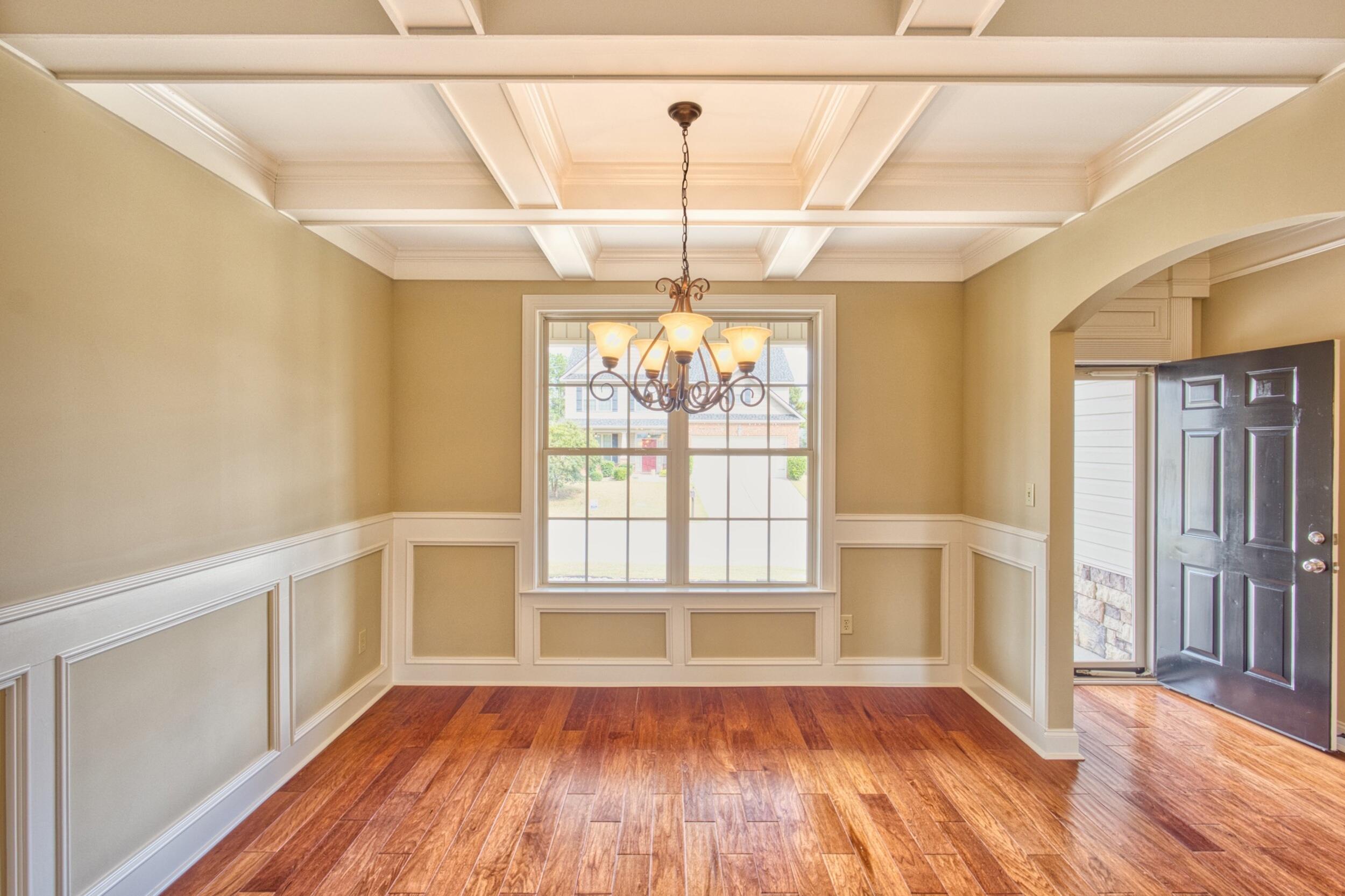 Empty dining room with a coffered ceiling, chandelier, wood floors and an open front door.