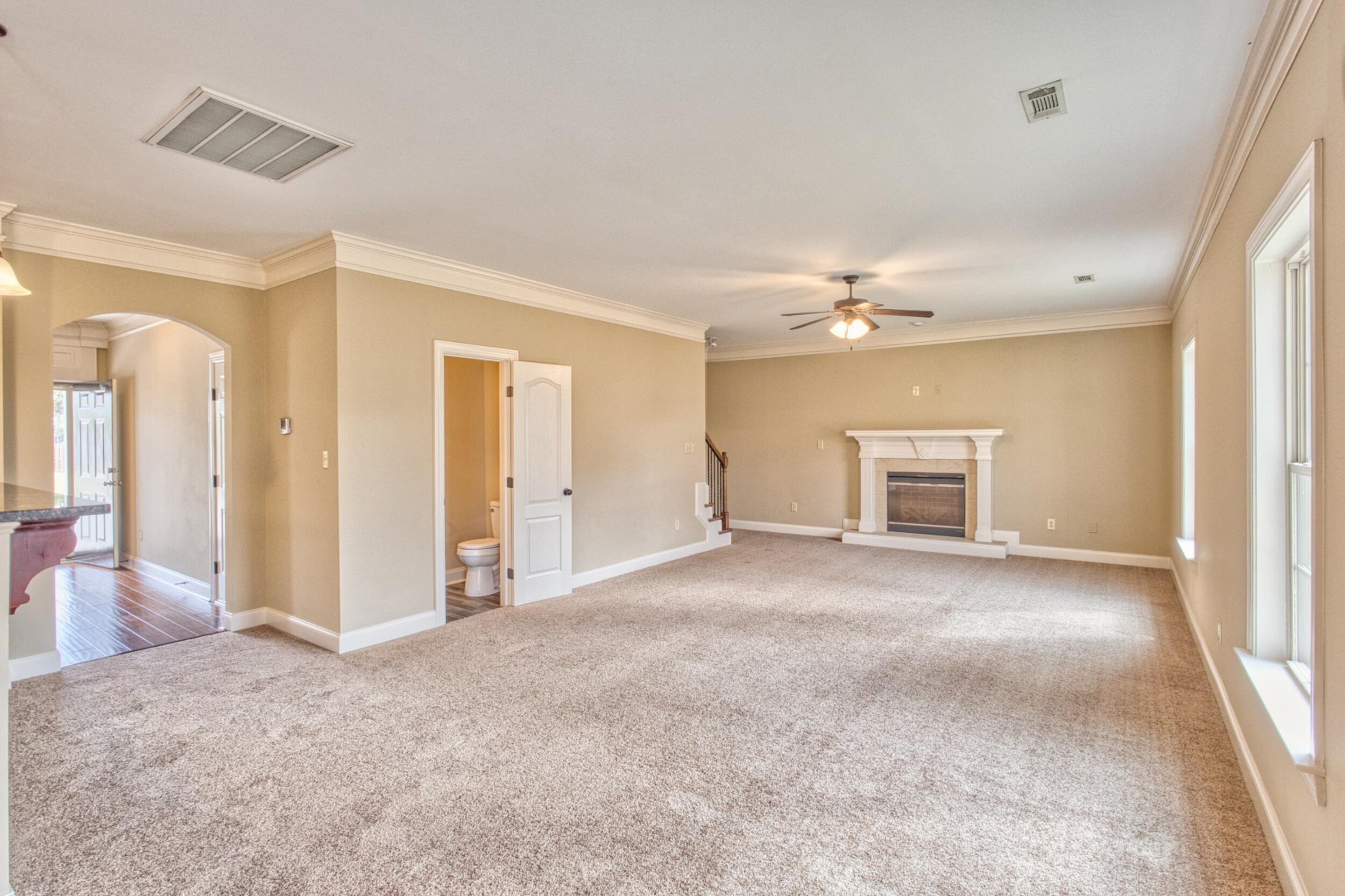 Vacant living room with a white-mantel fireplace, carpet, ceiling fan, and tall windows.
