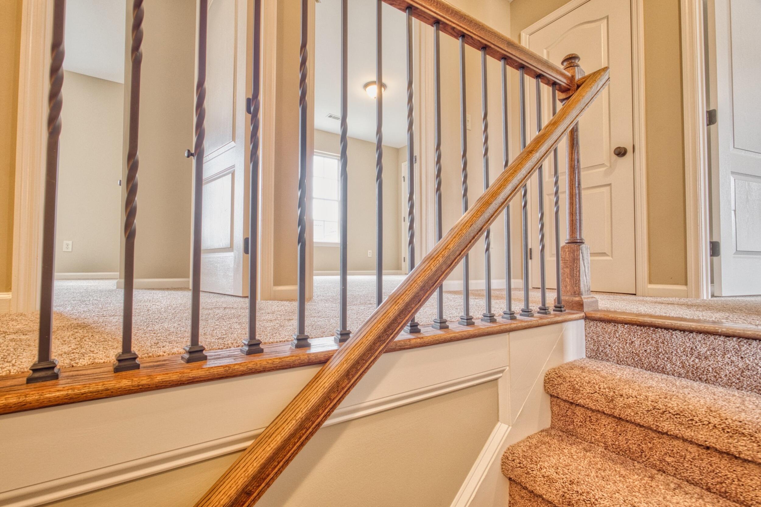 Carpeted staircase with a polished wood handrail and dark metal balusters leading to a bright upstairs landing.