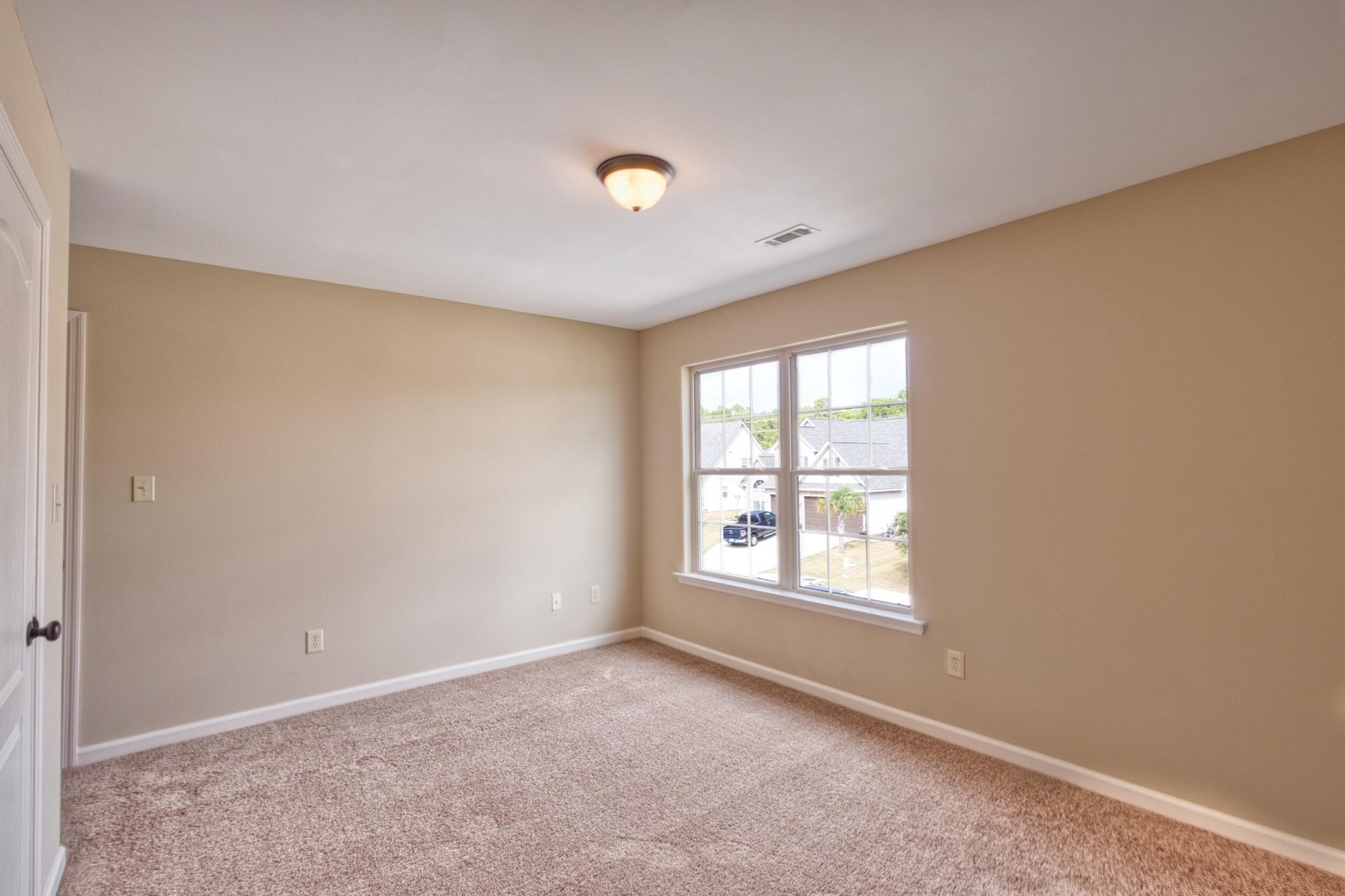 Empty carpeted bedroom with beige walls and a large double window overlooking neighboring homes.