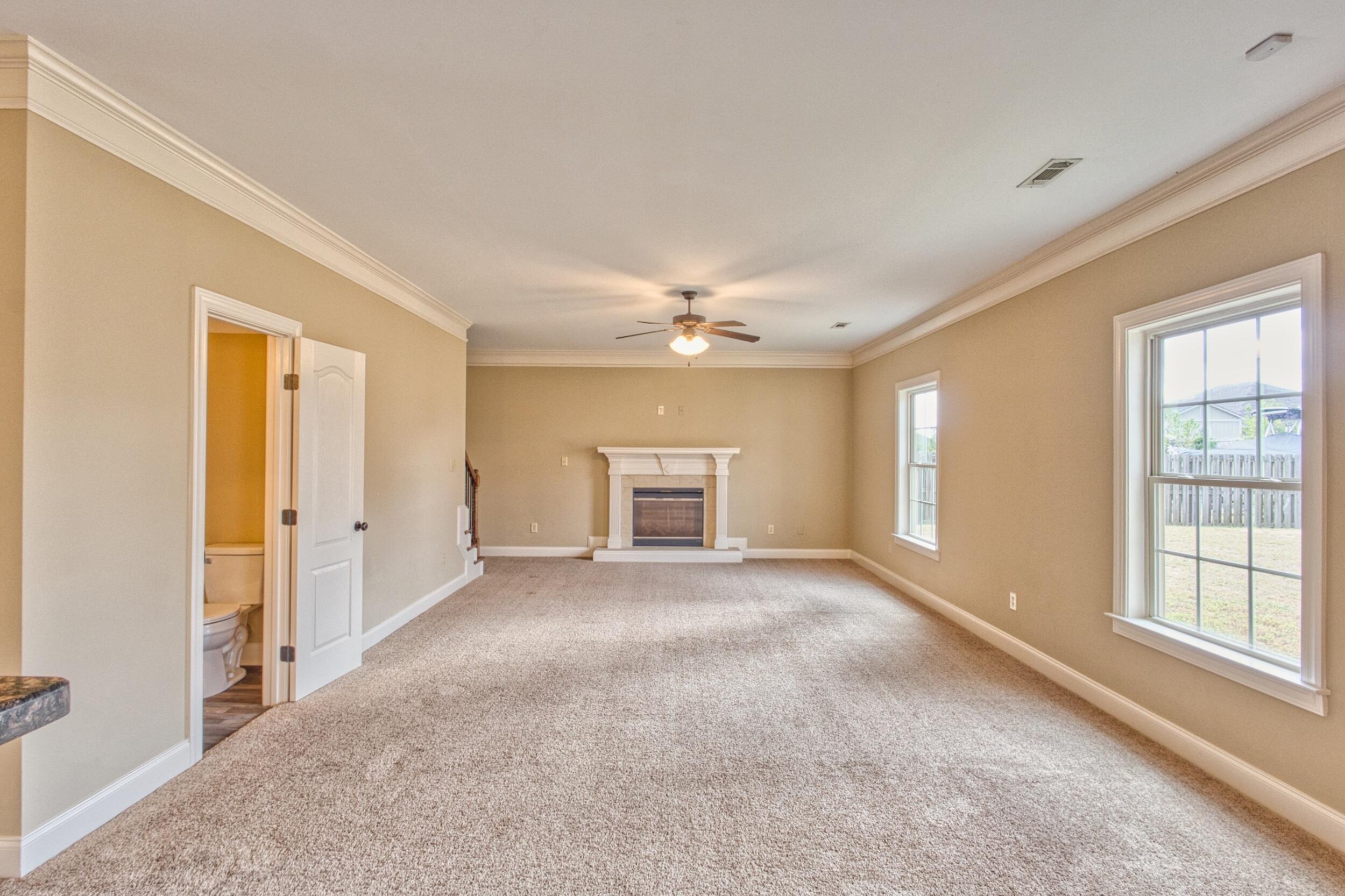 Unfurnished living room with a white fireplace, ceiling fan, and large windows overlooking a fenced yard.