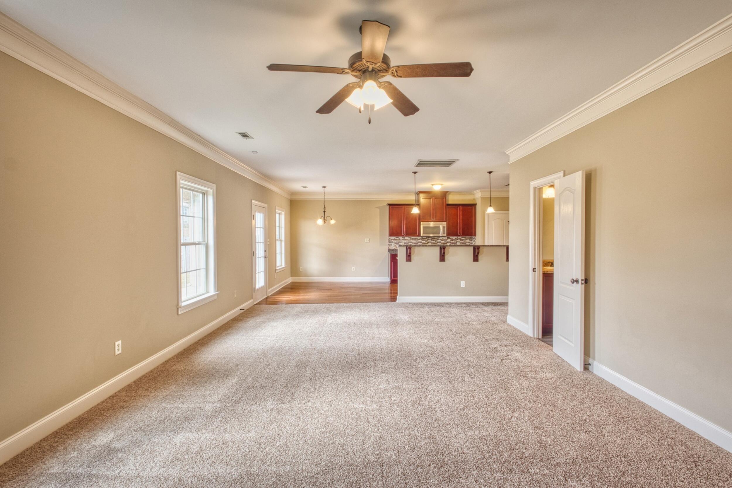 Empty carpeted living room opening to a dining area and kitchen with dark wood cabinets.