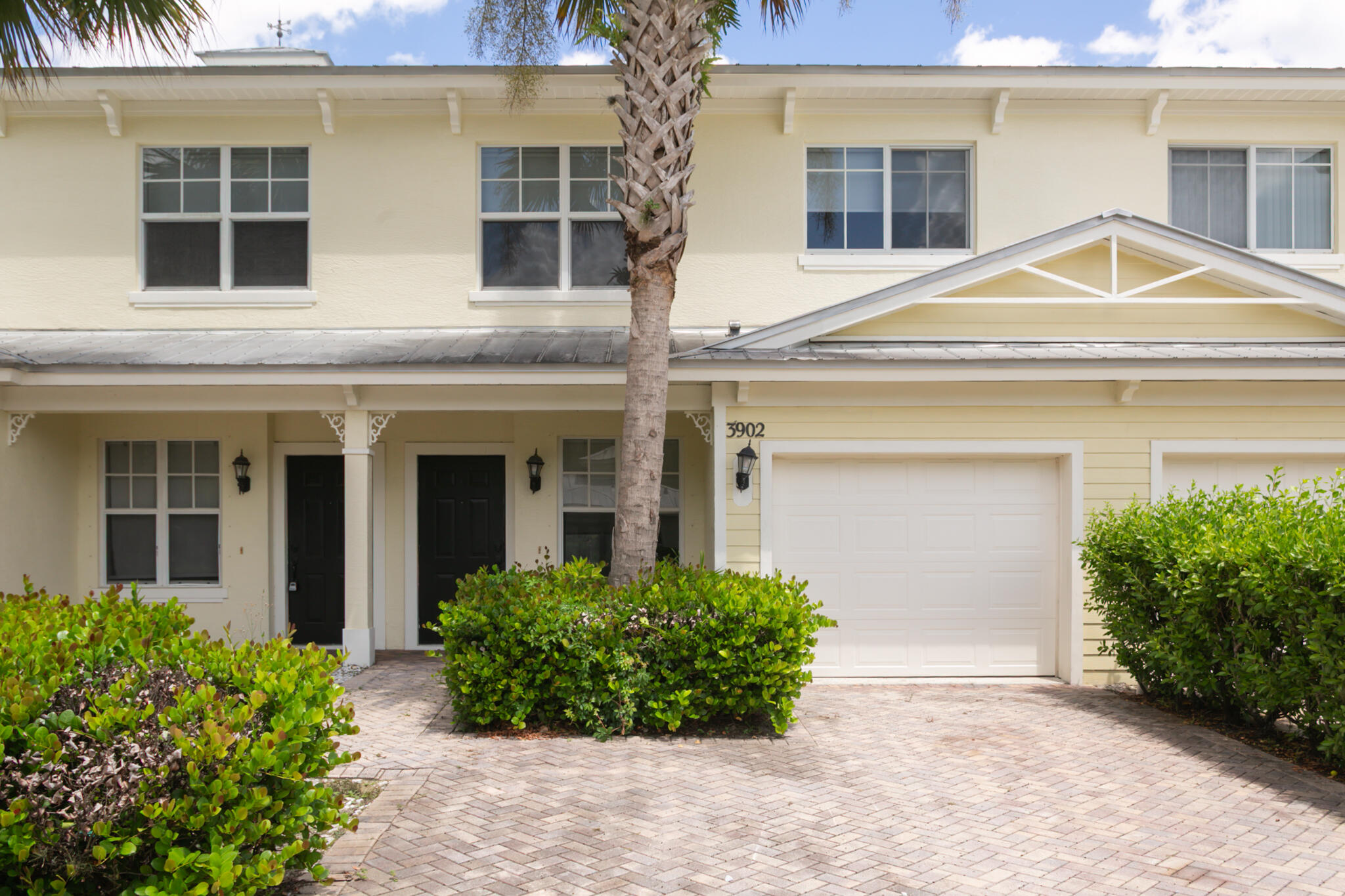Two-story home with a palm tree, trimmed shrubs, and a brick-paver driveway.