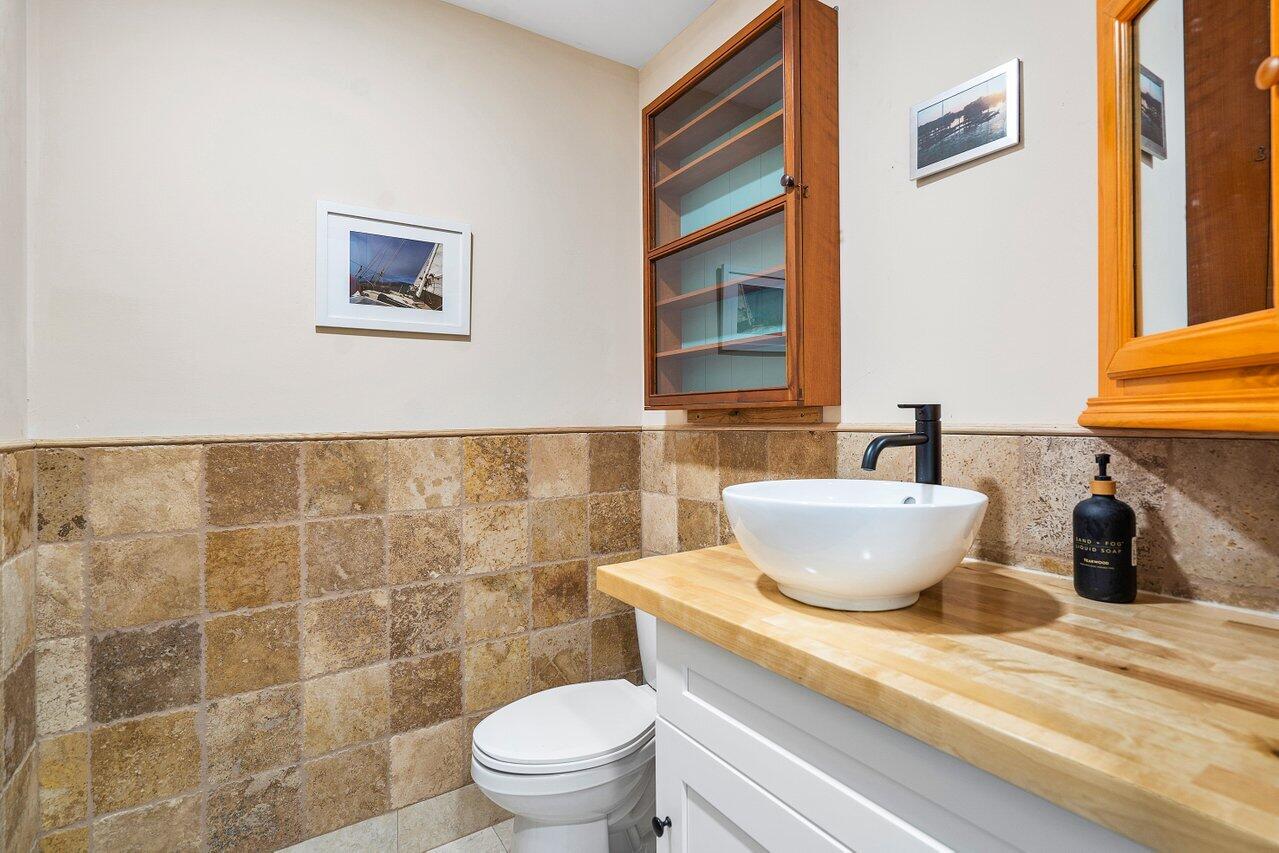 Bathroom with a vessel sink, light wood countertop, tan stone tile, and glass-front wood cabinet.