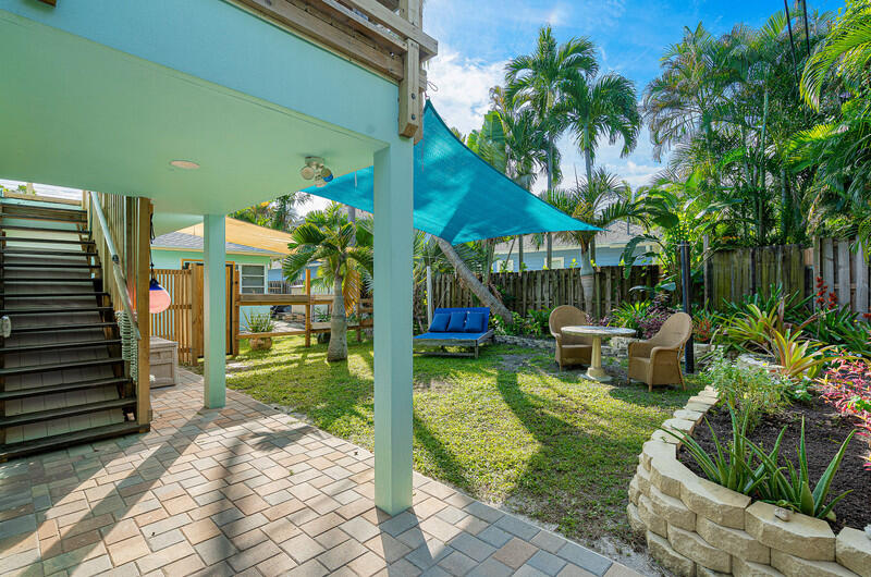 Backyard patio with a blue shade sail, outdoor seating, tropical plants, and stairs beside the house.