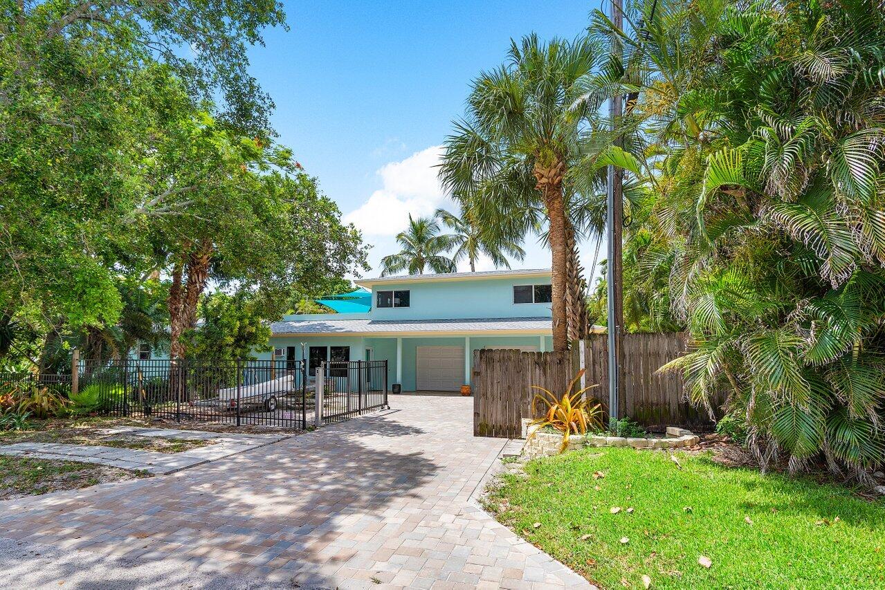 A pale aqua home behind a paved driveway, framed by palms and a gated entrance.